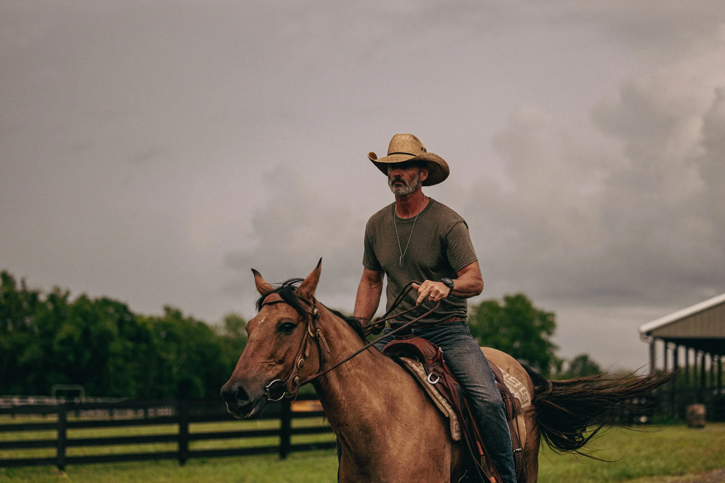A man riding a horse outdoors on a cloudy day, wearing a cowboy hat and casual clothing.