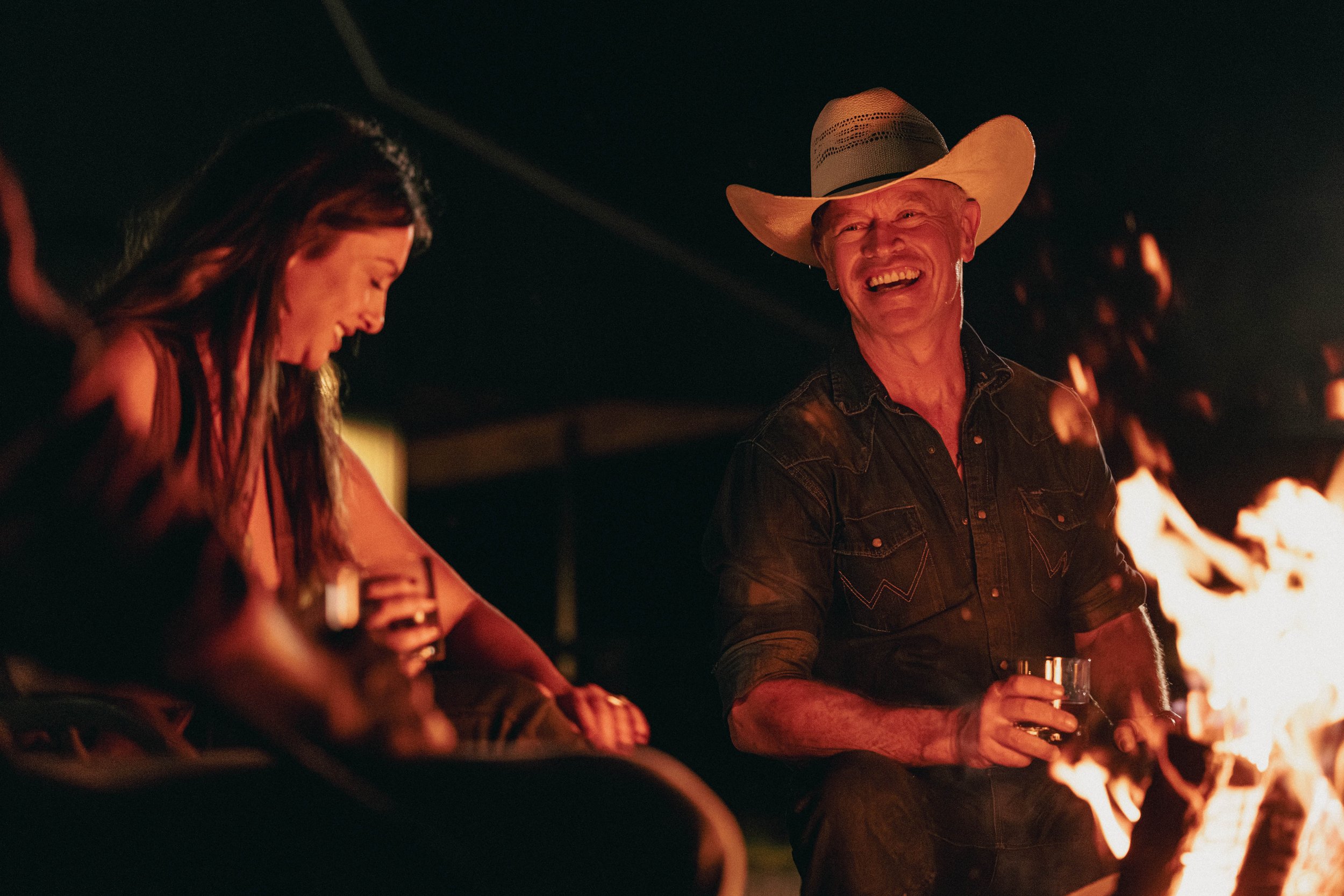 Two people sitting around a campfire at night, laughing and holding drinks, with the man wearing a cowboy hat.