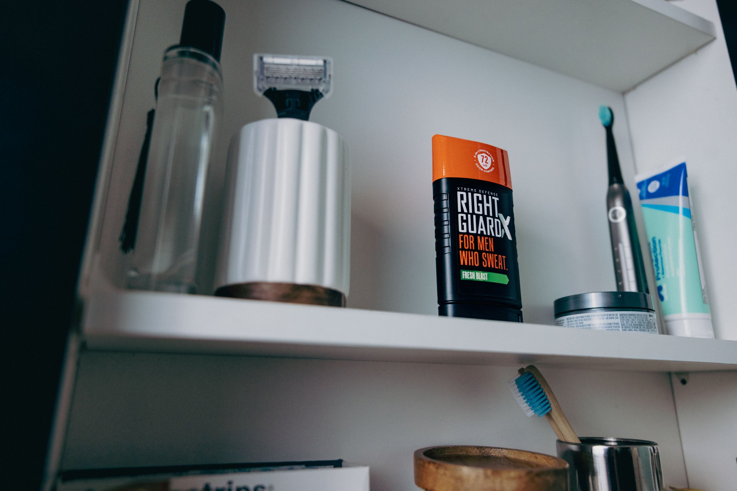 Bathroom shelf containing a spray bottle, an electric trimmer, a can of Right Guard deodorant, a toothbrush, toothpaste, and a small container.