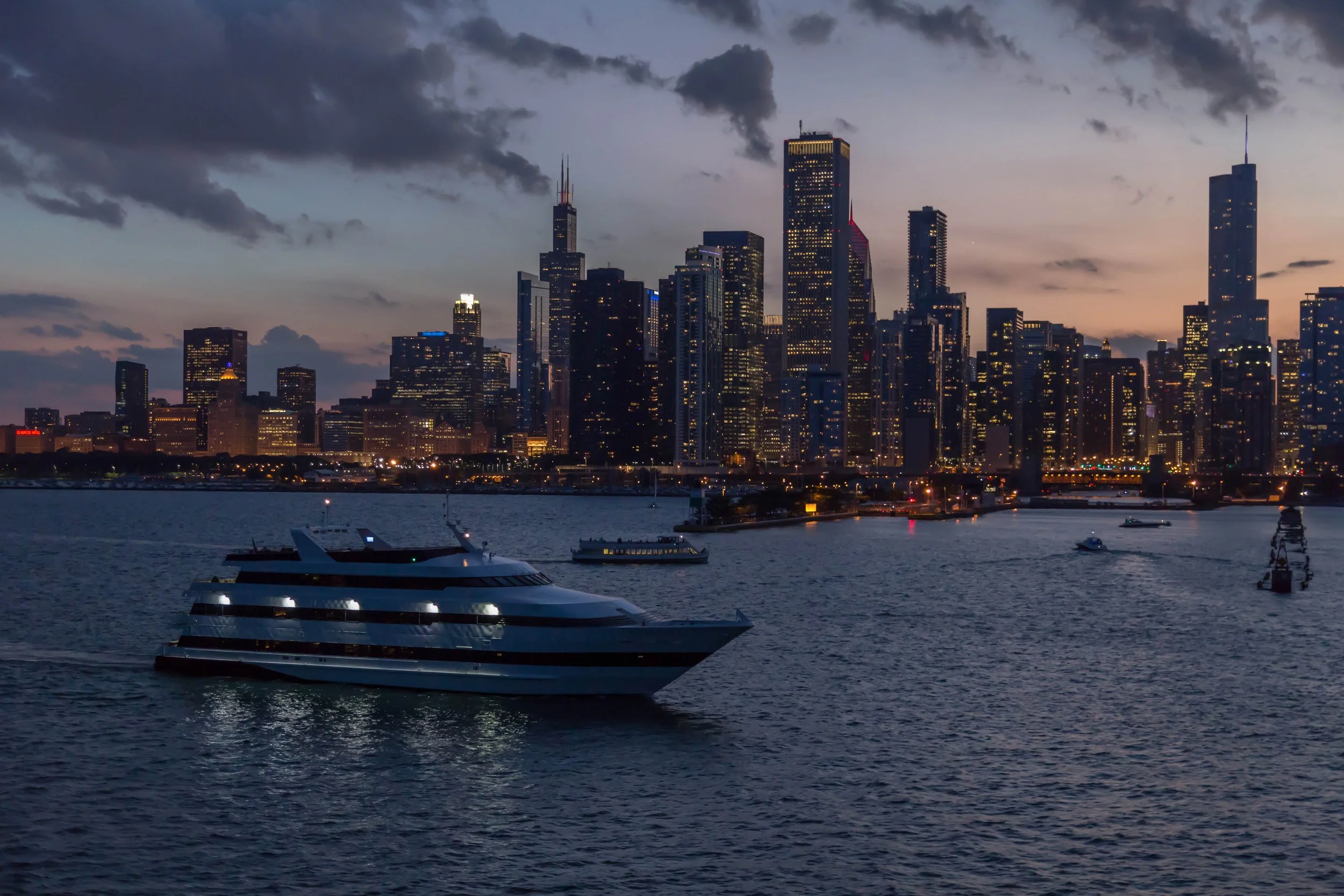 Skyline of a city with tall modern buildings and skyscrapers during dusk, with dark clouds in the sky and water with boats in the foreground.