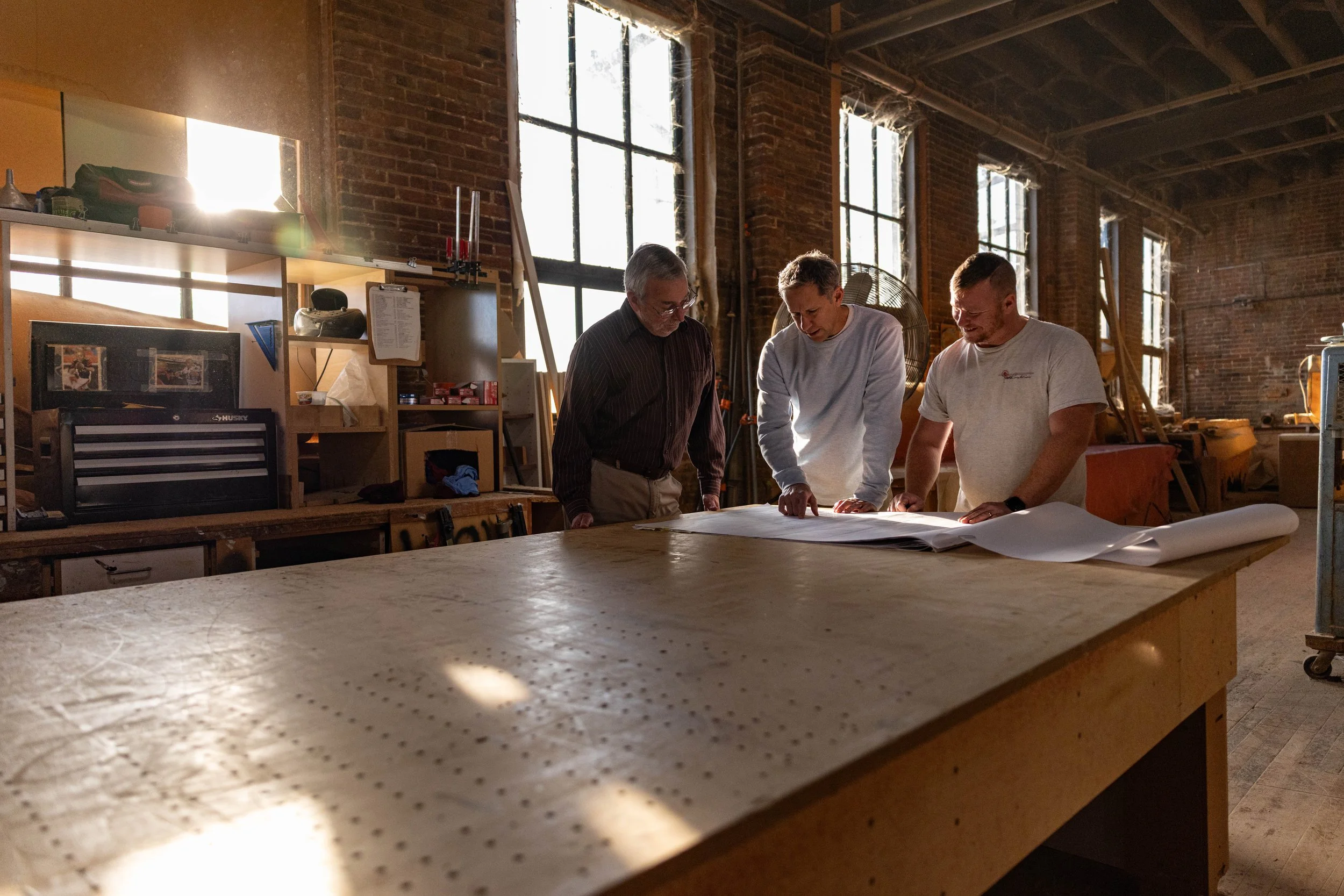 Three men in a workshop examining blueprints on a large wooden table with high windows and brick walls in the background.
