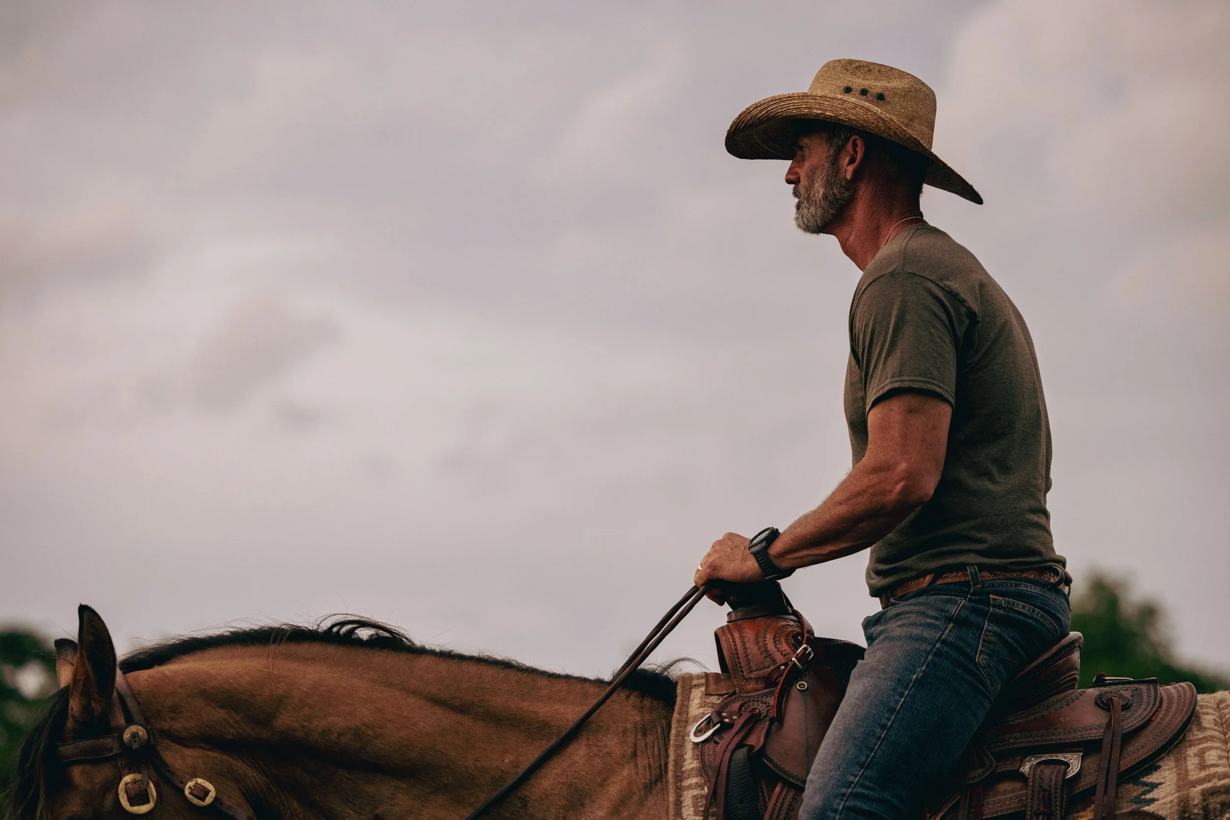 A man wearing a wide-brimmed hat, dark T-shirt, and jeans rides a brown horse outdoors on a cloudy day.