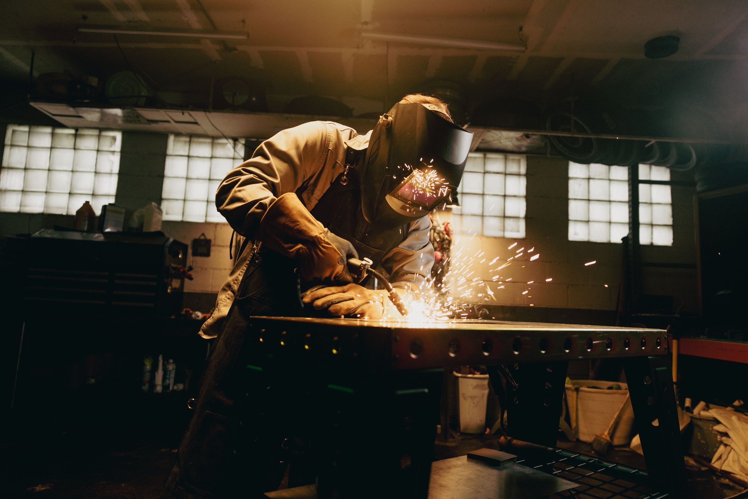 Person welding metal in a workshop, wearing a protective welding helmet and gloves, with sparks flying.