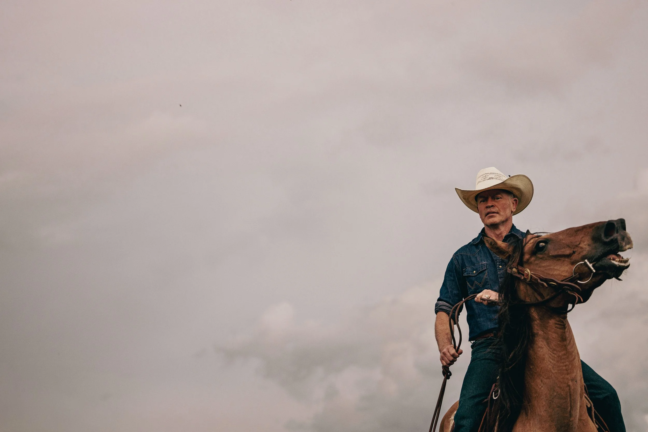 A man wearing a cowboy hat and denim shirt riding a brown horse against a cloudy sky.