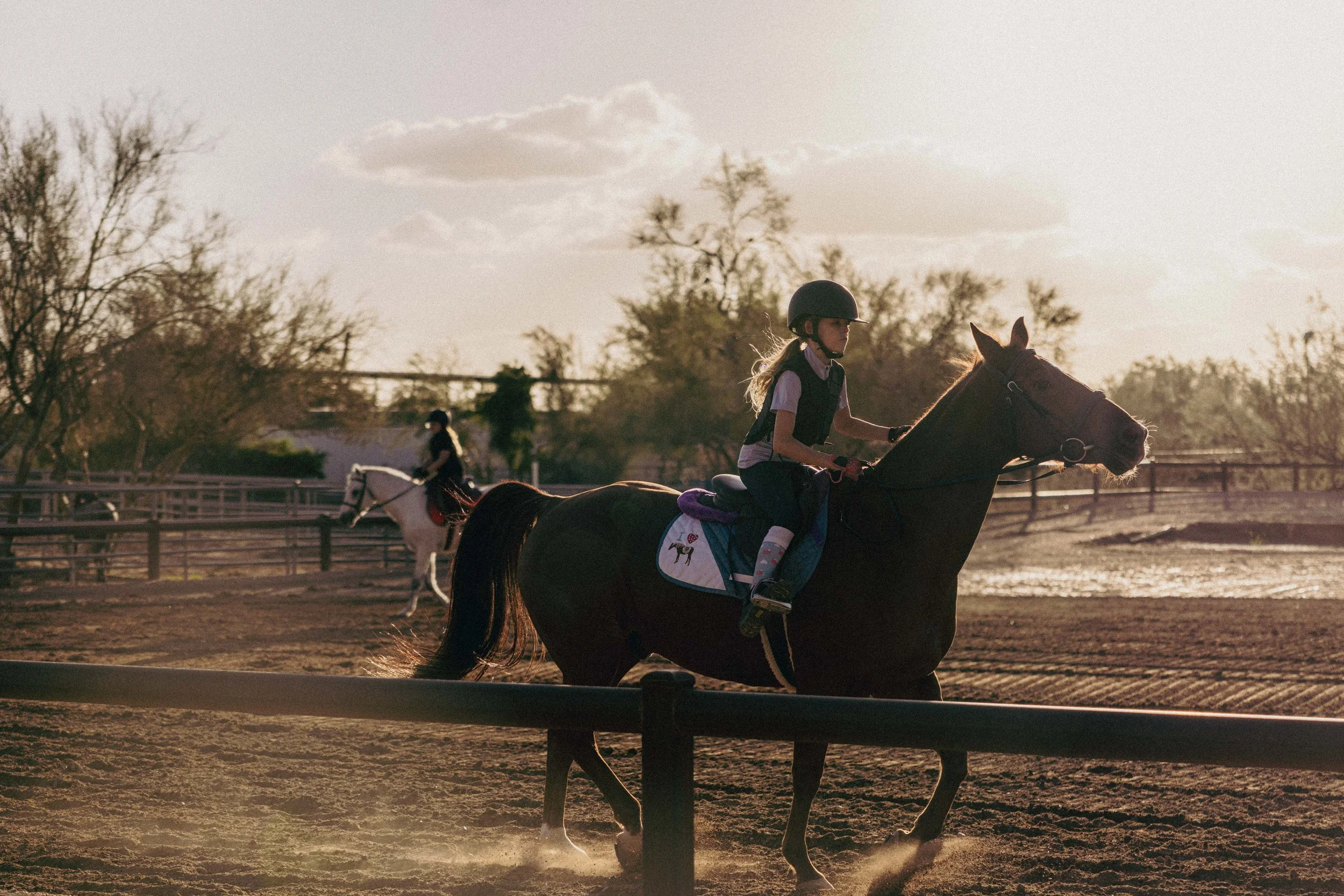 A young girl riding a horse on a dirt track during sunset, with another girl riding a white horse in the background.