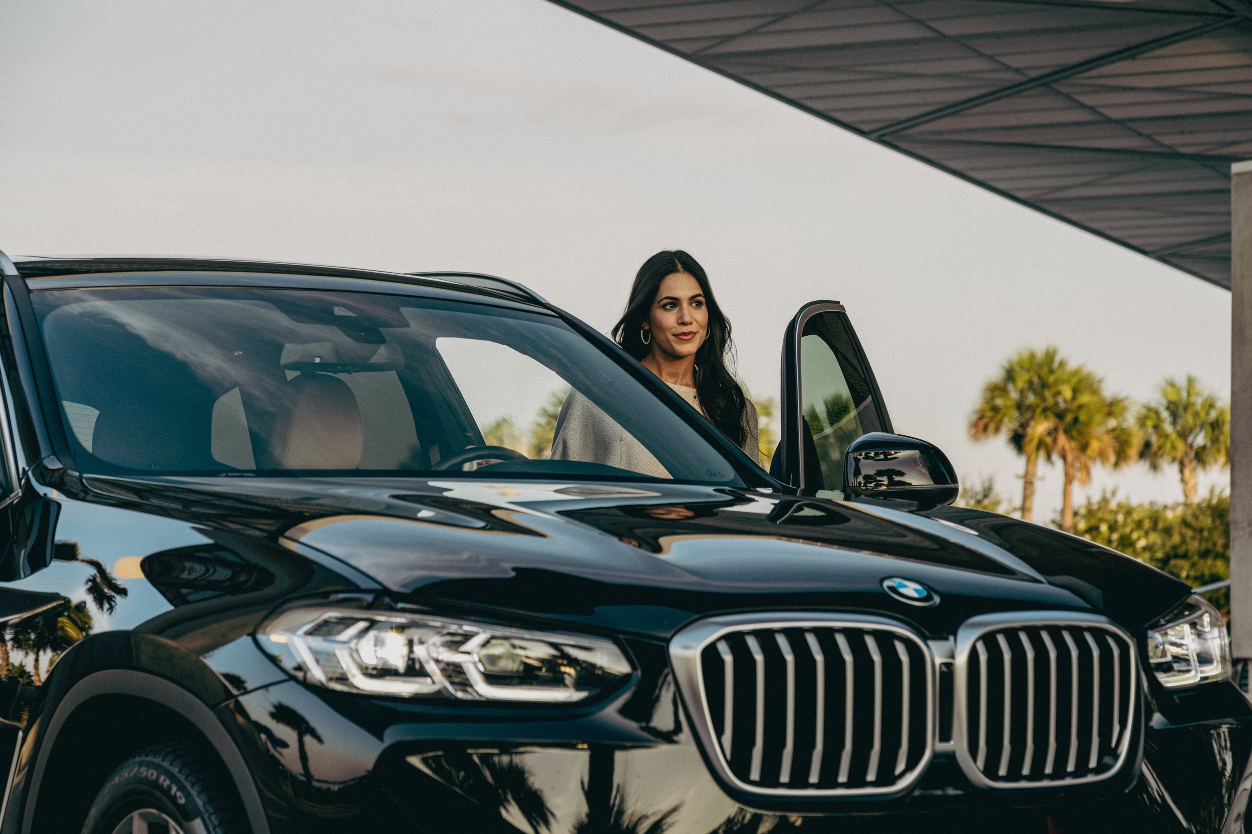 A woman with long dark hair standing next to a black BMW car with an open door, outdoors with trees and a building overhead.