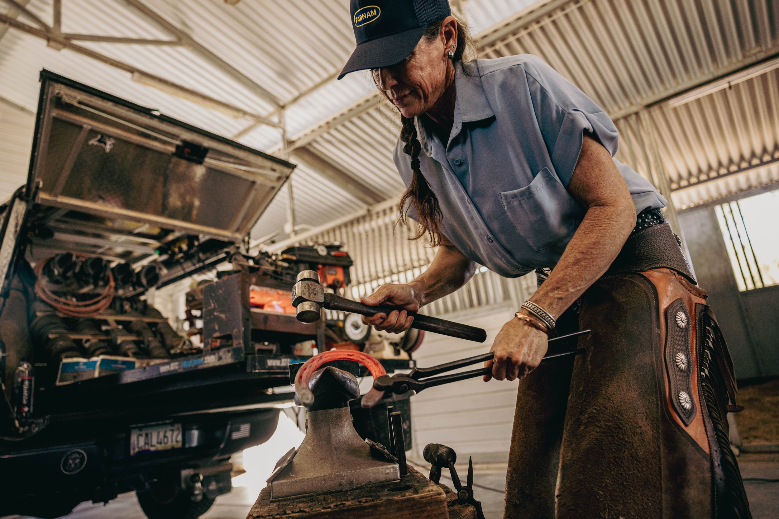 A woman wearing a mechanic's apron, cap, and jewelry is hammering a metal rod in a garage or workshop, with a vehicle open and tools on a workbench in the background.