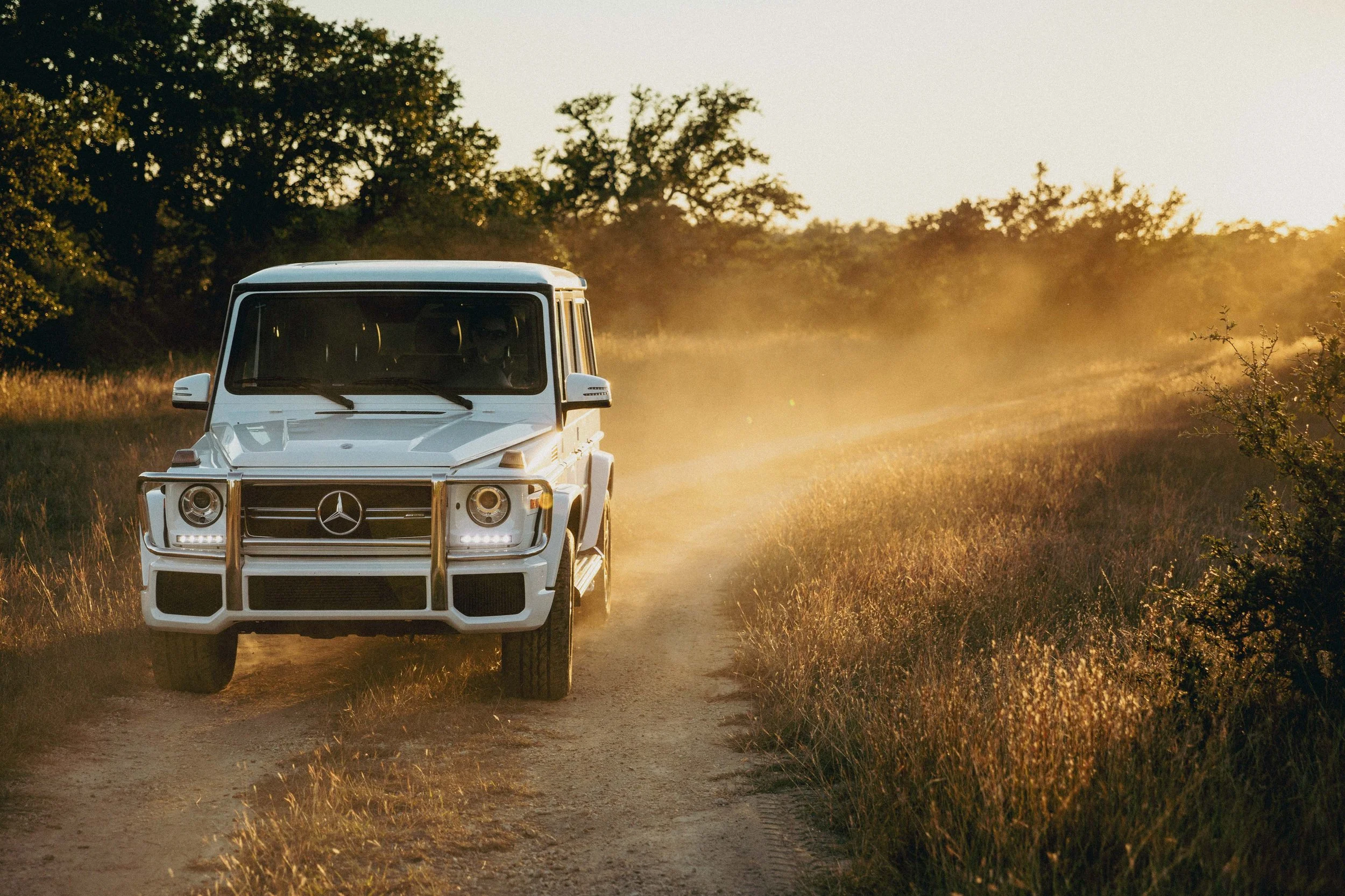 A white Mercedes-Benz G-Class SUV driving on a dirt road through a grassy landscape at sunset.