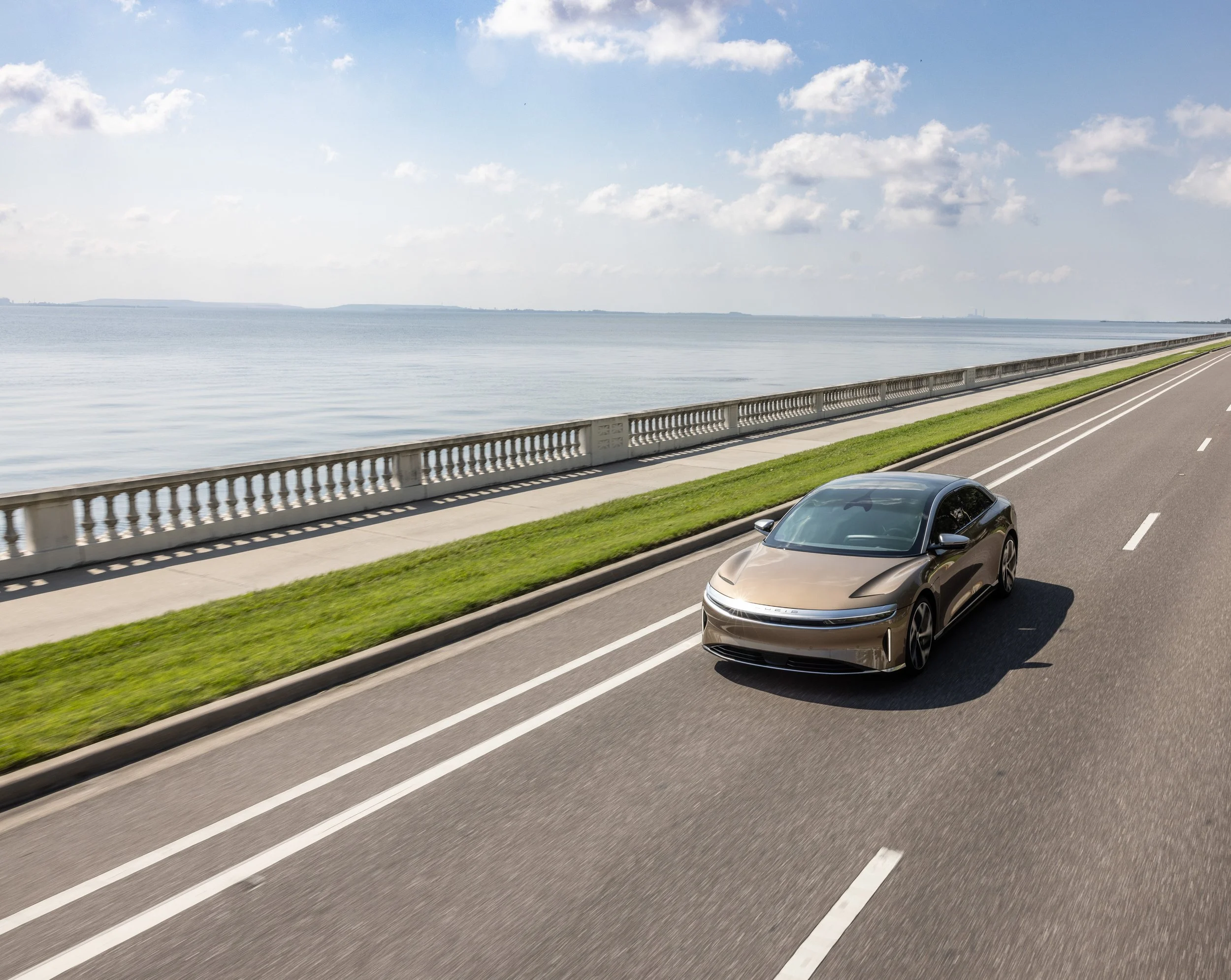 A beige car driving along a coastal road with a body of water and a partly cloudy sky in the background.