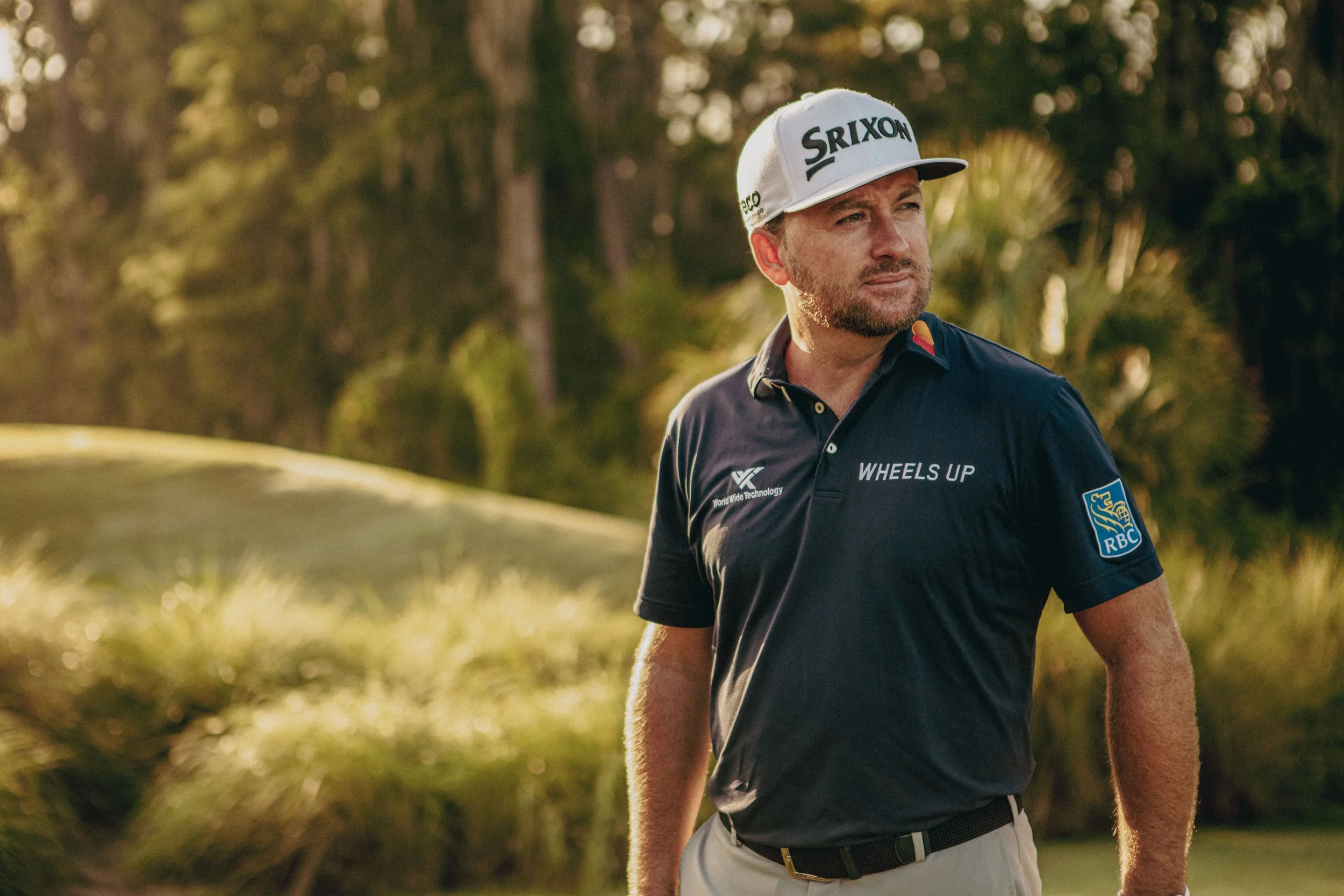 A man wearing a white cap and a navy blue polo shirt with logos, standing outdoors with trees and foliage in the background, looking to his right.