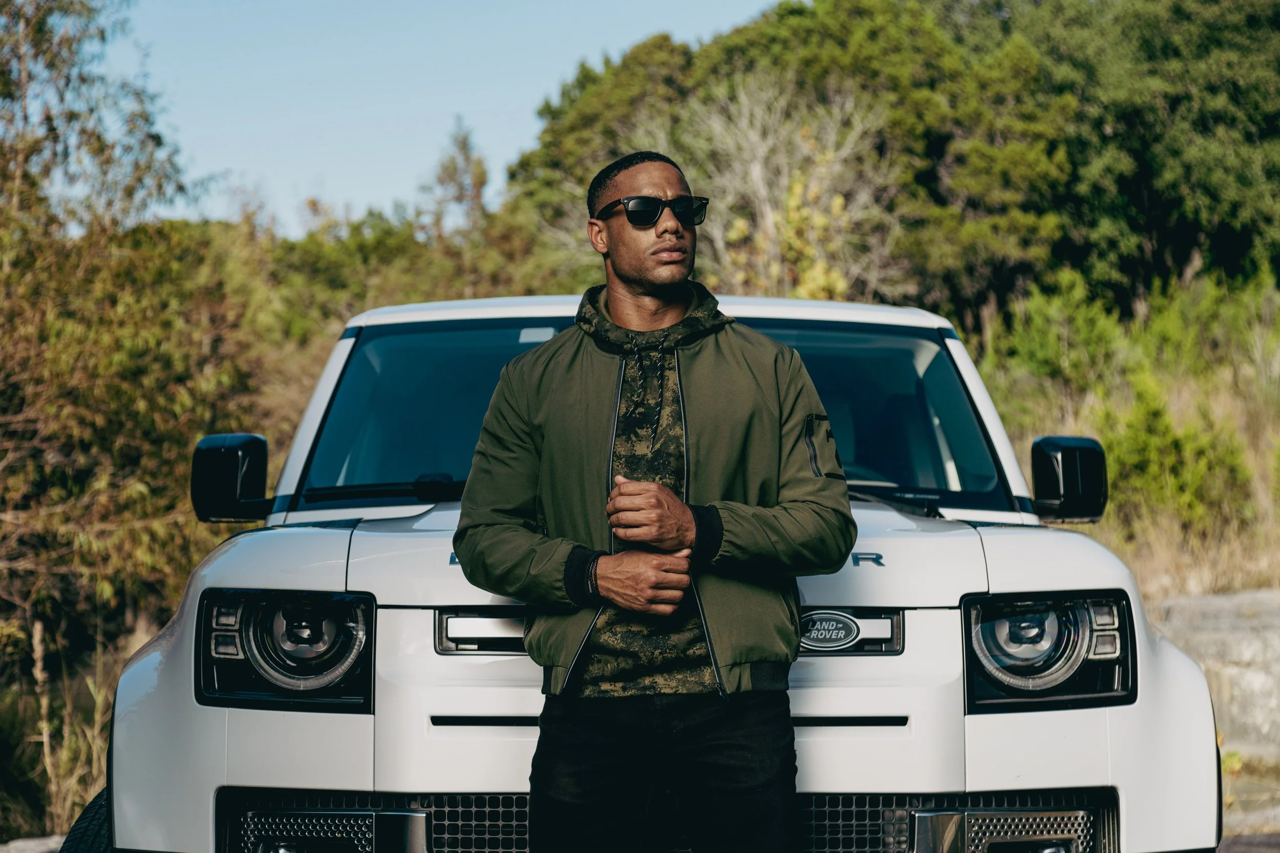 A man wearing sunglasses and a green jacket standing in front of a white Land Rover vehicle outdoors with trees and clear sky in the background.