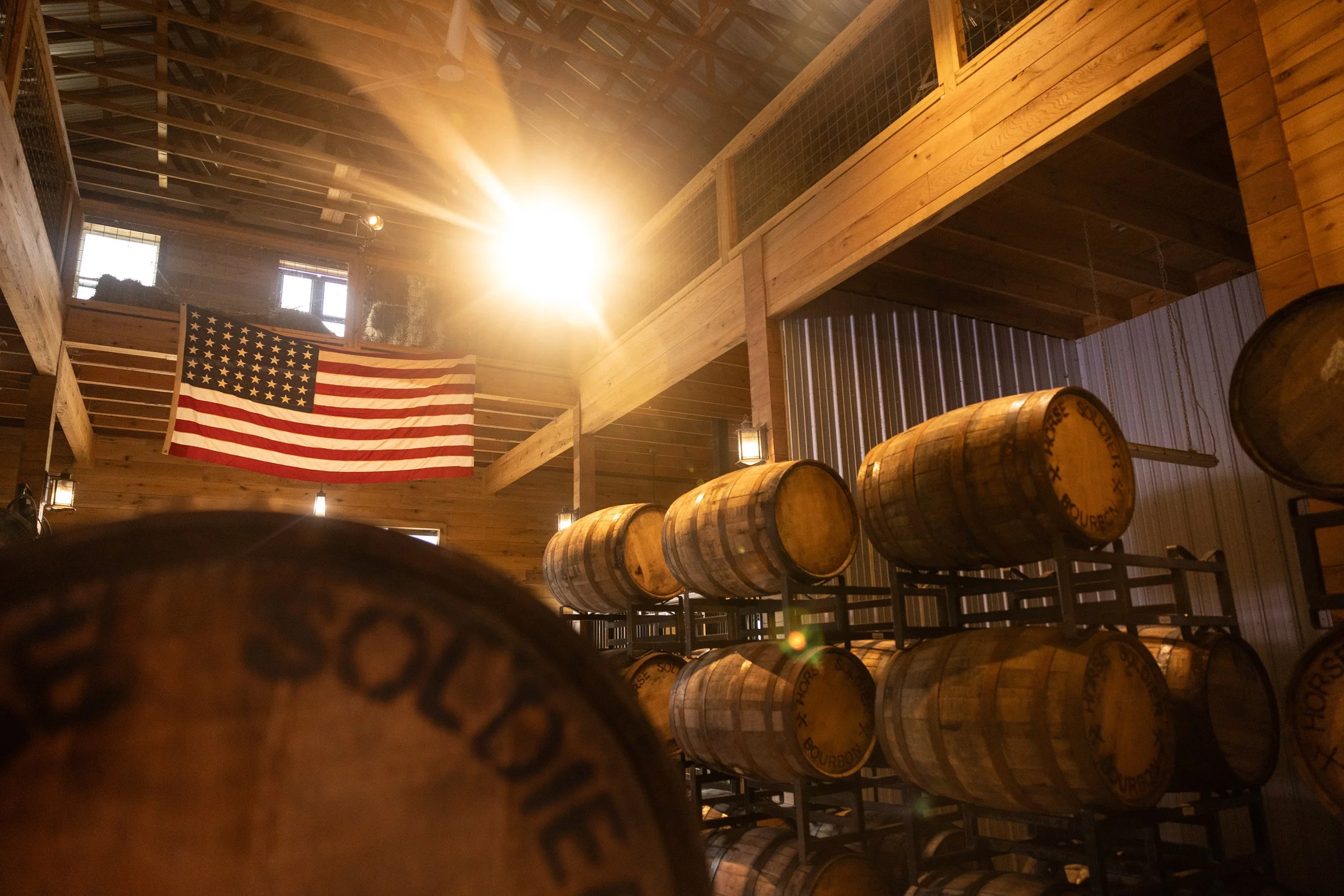 Wooden barrels stored in a distillery, with an American flag hanging on the wall and warm sunlight coming through the windows.