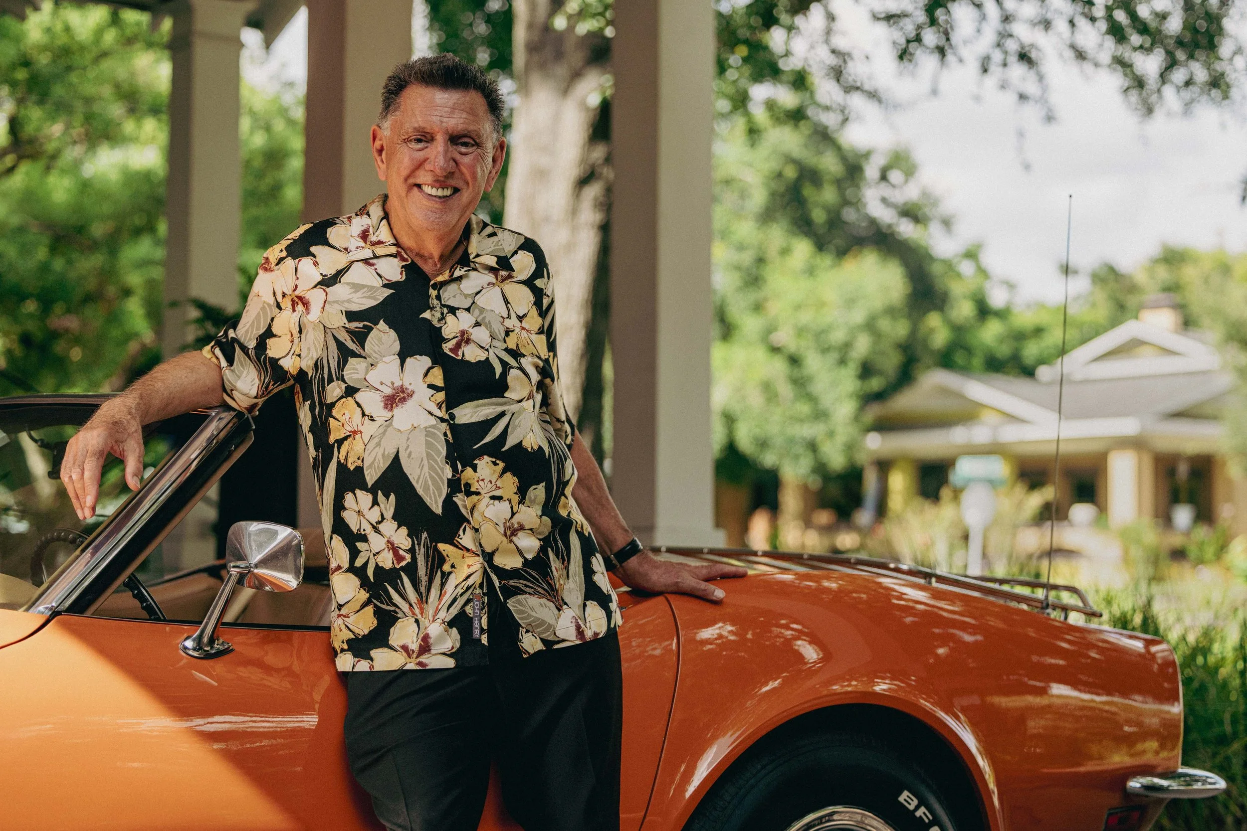 An elderly man with gray hair smiling, wearing a floral shirt, standing beside an orange vintage car on a porch with trees and a house in the background.