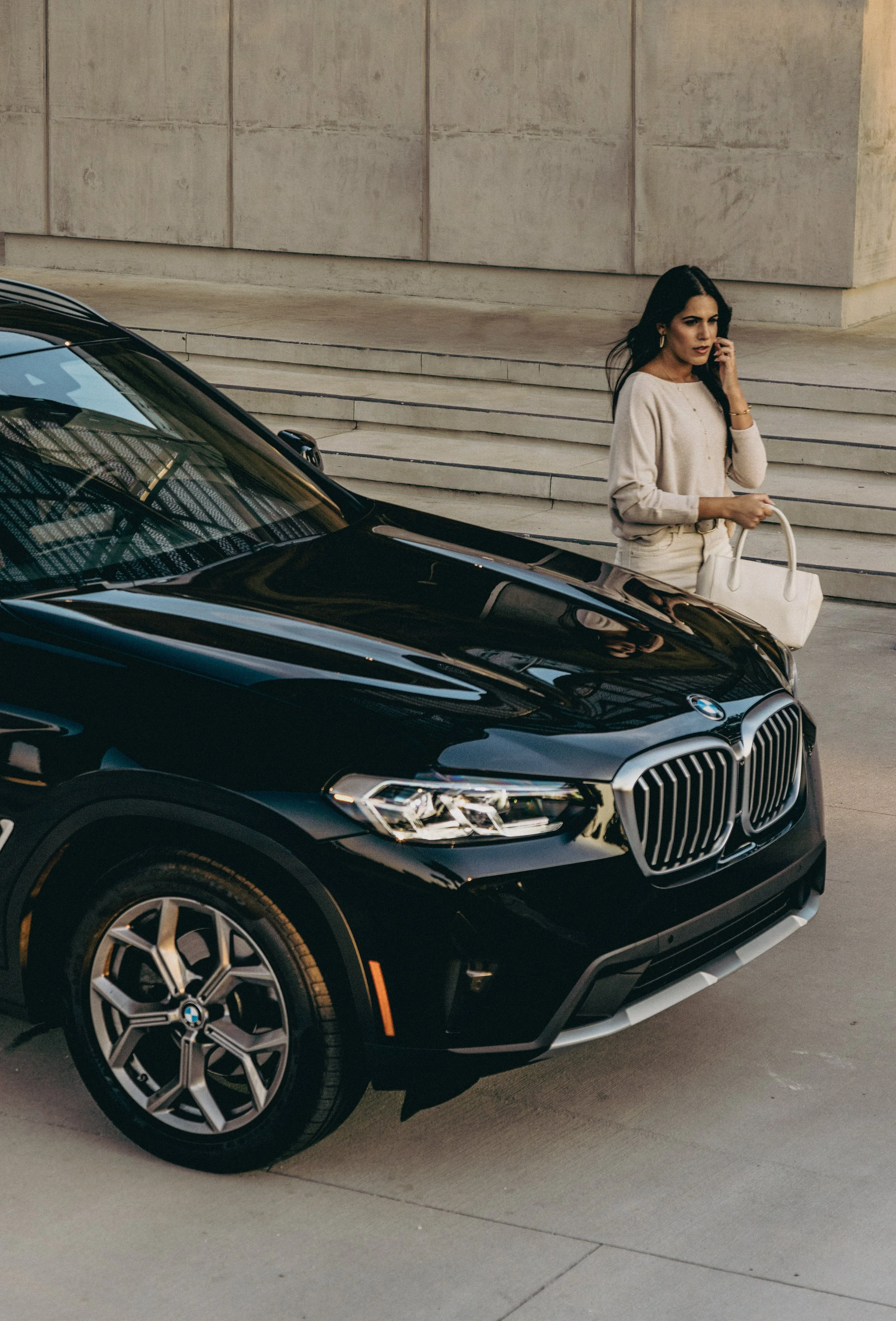 A woman standing next to a black BMW SUV, talking on her cellphone, holding a white handbag, in front of a building with steps.