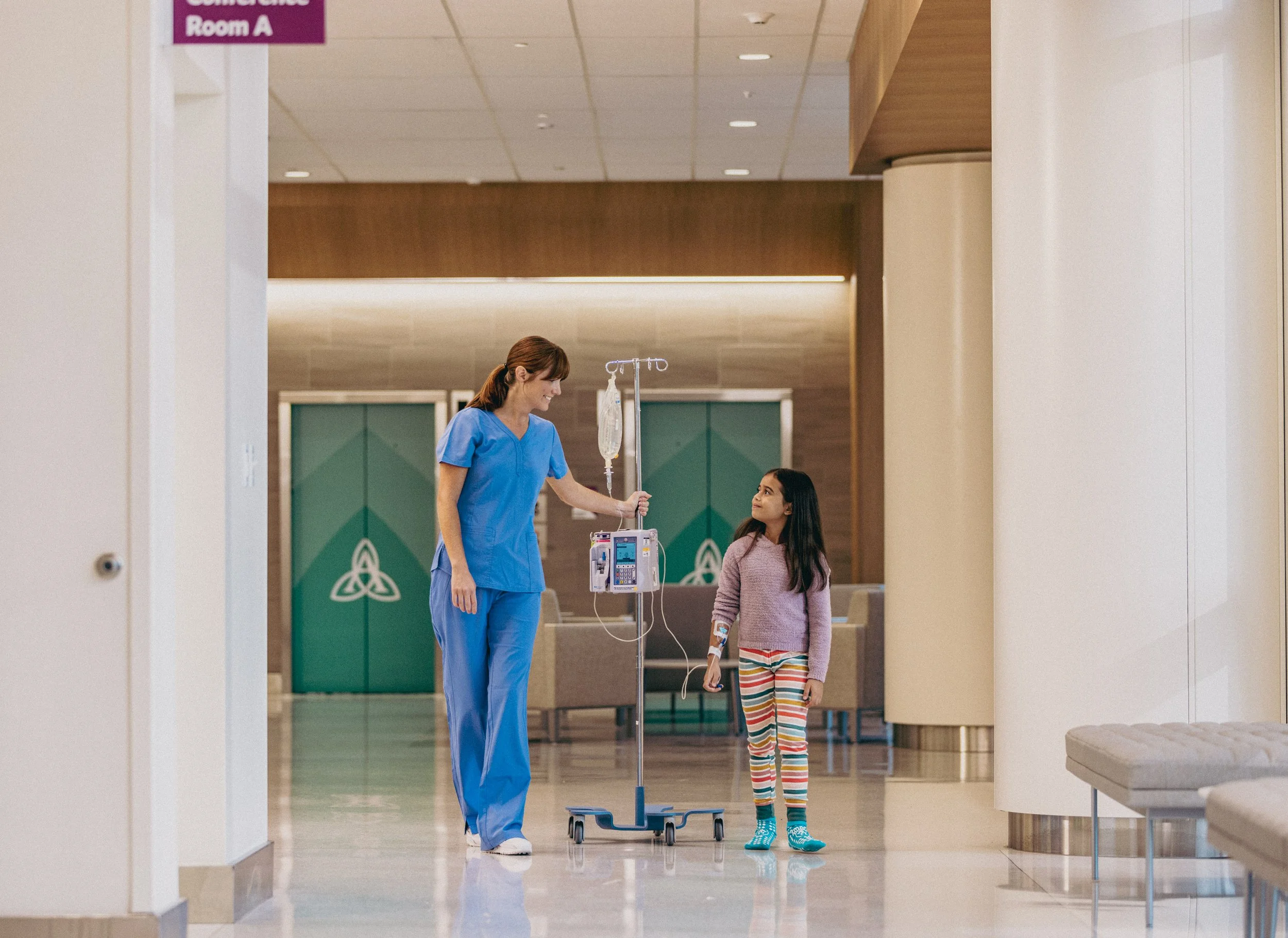 A healthcare worker, dressed in blue scrubs, holding an IV pole with an IV bag, smiling and talking to a young girl with a medical bandage on her arm in a hospital lobby.