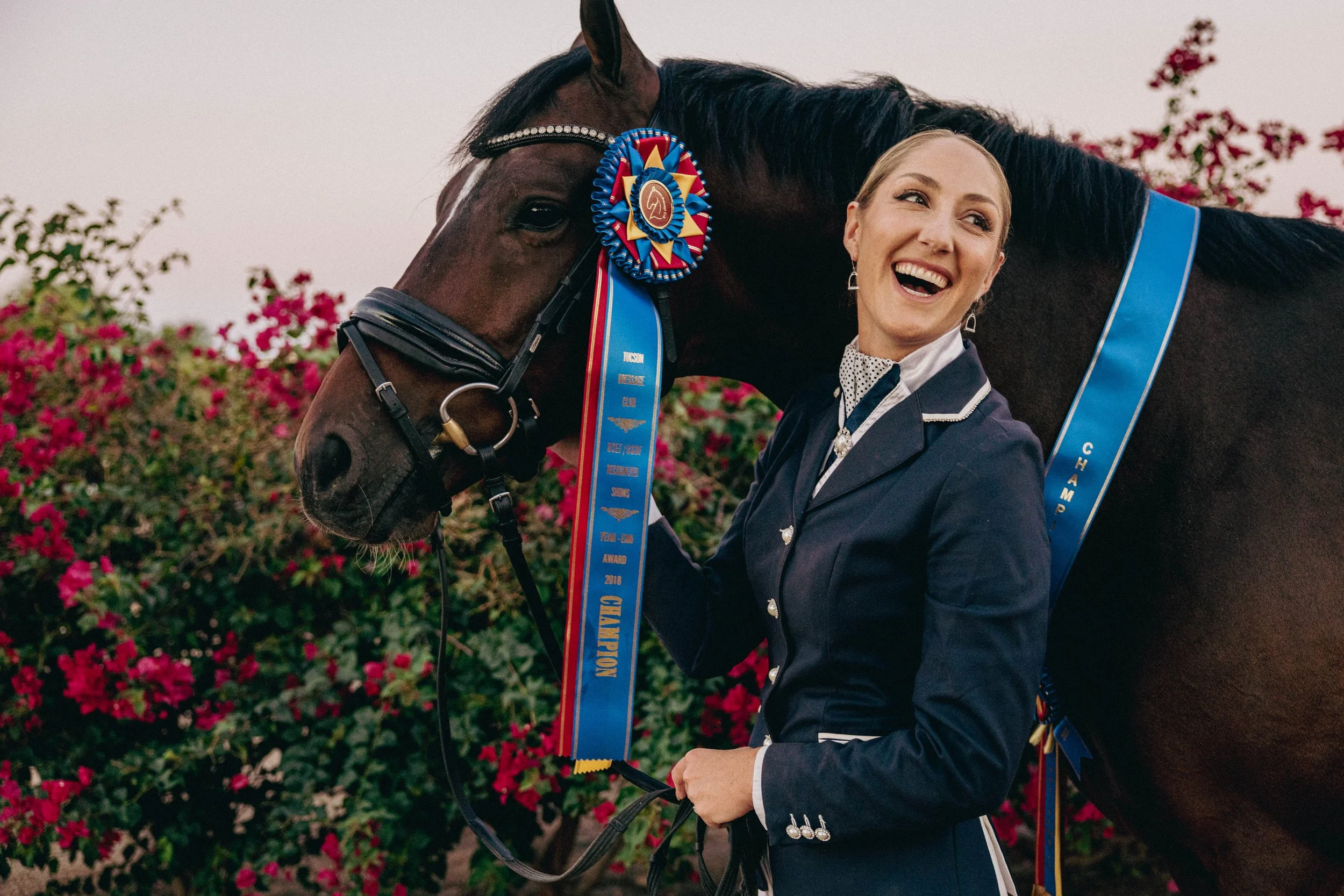 A woman dressed in an equestrian outfit smiling and holding the bridle of a dark brown horse with a blue and red ribbon rosette. The horse is adorned with a blue sash with gold lettering and embroidery. Pink flowering bushes are in the background.
