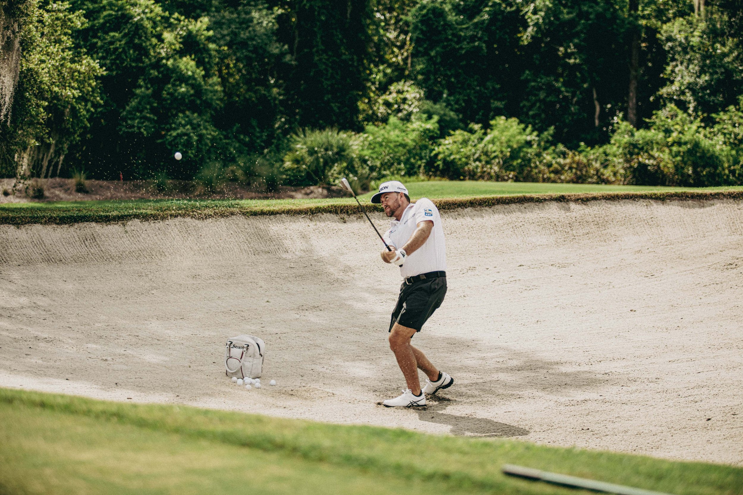 A golfer hitting a shot from a sand bunker onto a golf course, surrounded by green grass and trees.