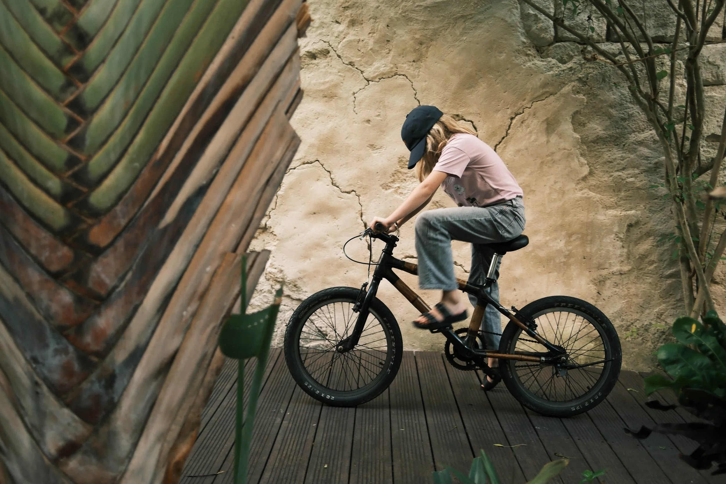 A young girl riding a black bicycle on a wooden deck against a textured, cracked wall, with green plants nearby.