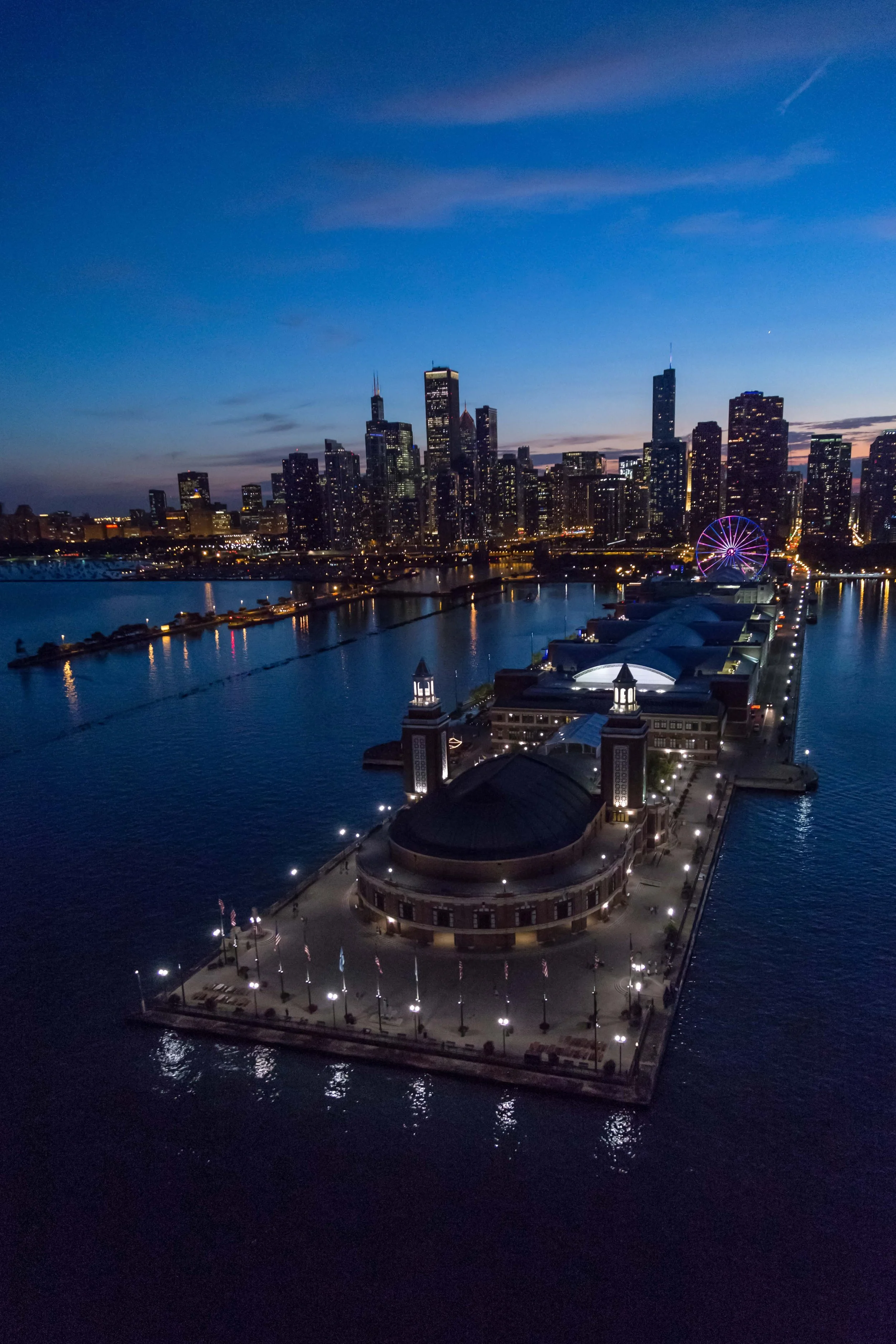 Nighttime aerial view of Navy Pier in Chicago with the city skyline in the background, illuminated Ferris wheel, and calm water reflections.