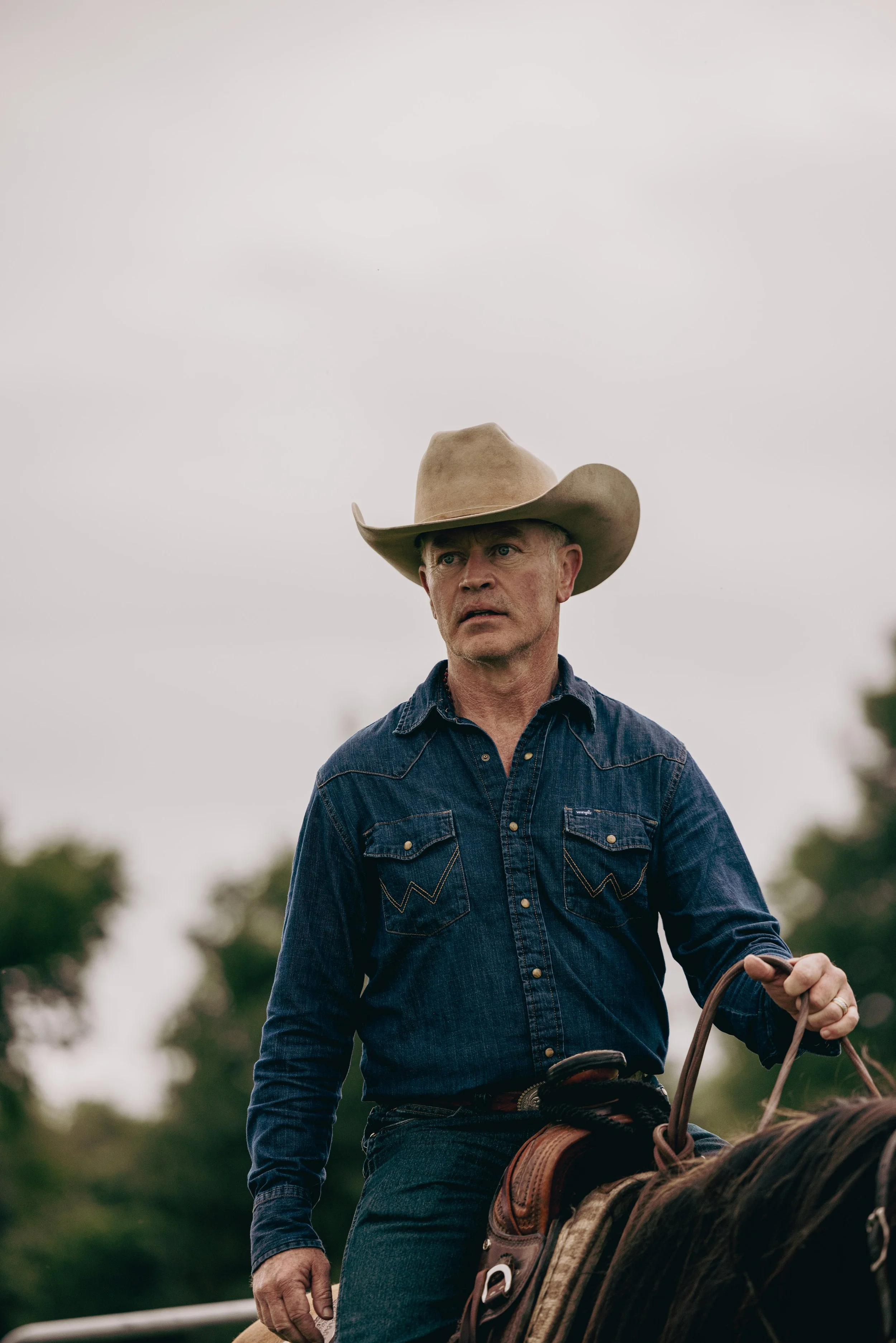 A man wearing a cowboy hat and denim shirt riding a horse outdoors.