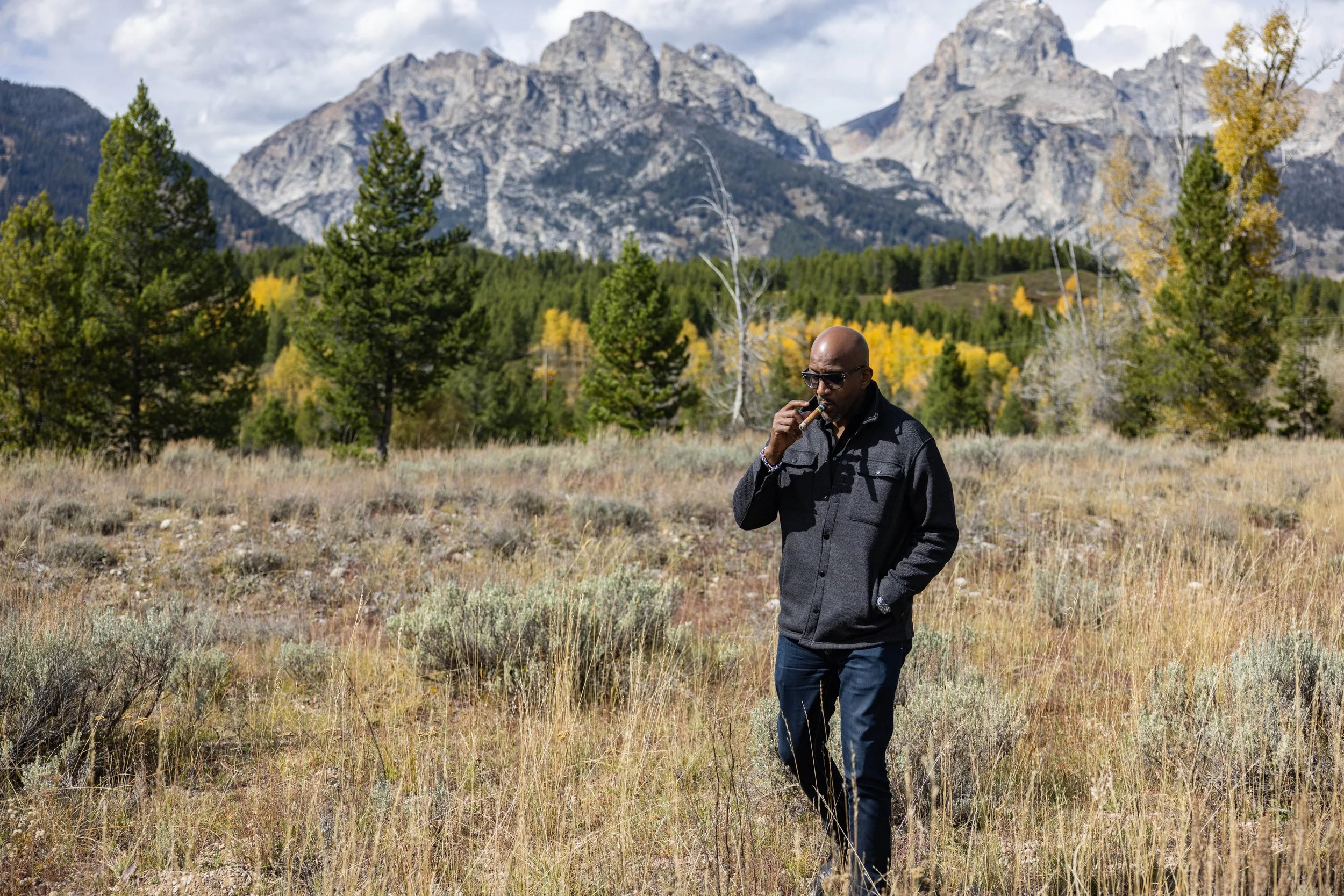 A man with sunglasses in a black jacket walking through a field of dry grass, with a forest, mountains, and cloudy sky in the background.