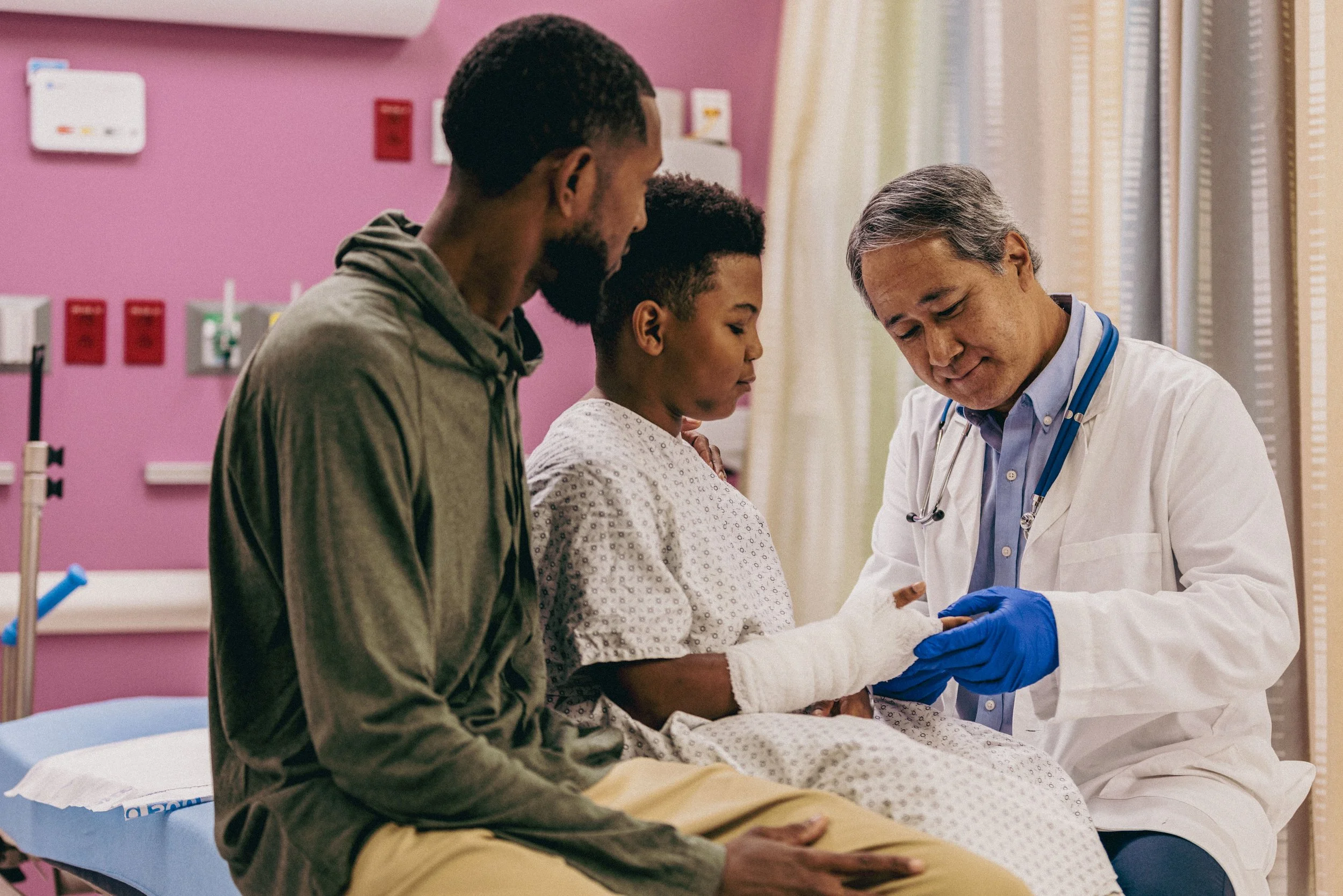 A doctor in a white coat and blue gloves examining a young girl in a hospital gown, with her father and mother sitting beside her.