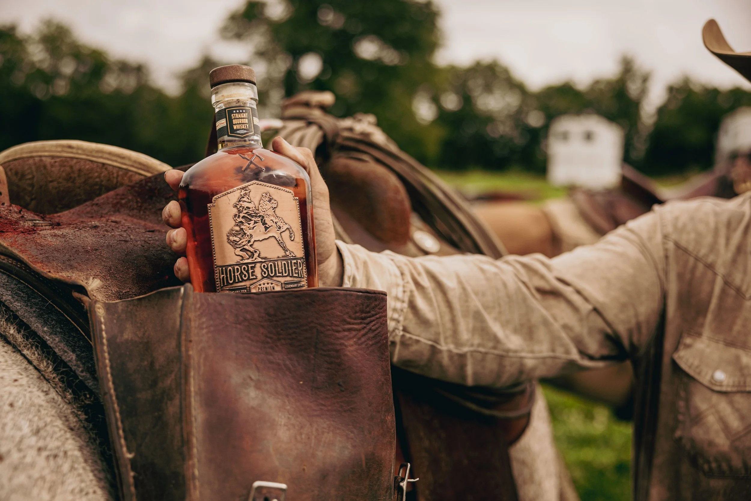 Person holding a bottle of Horse Soldier bourbon next to a saddle on a horse, outdoors in a rural setting.