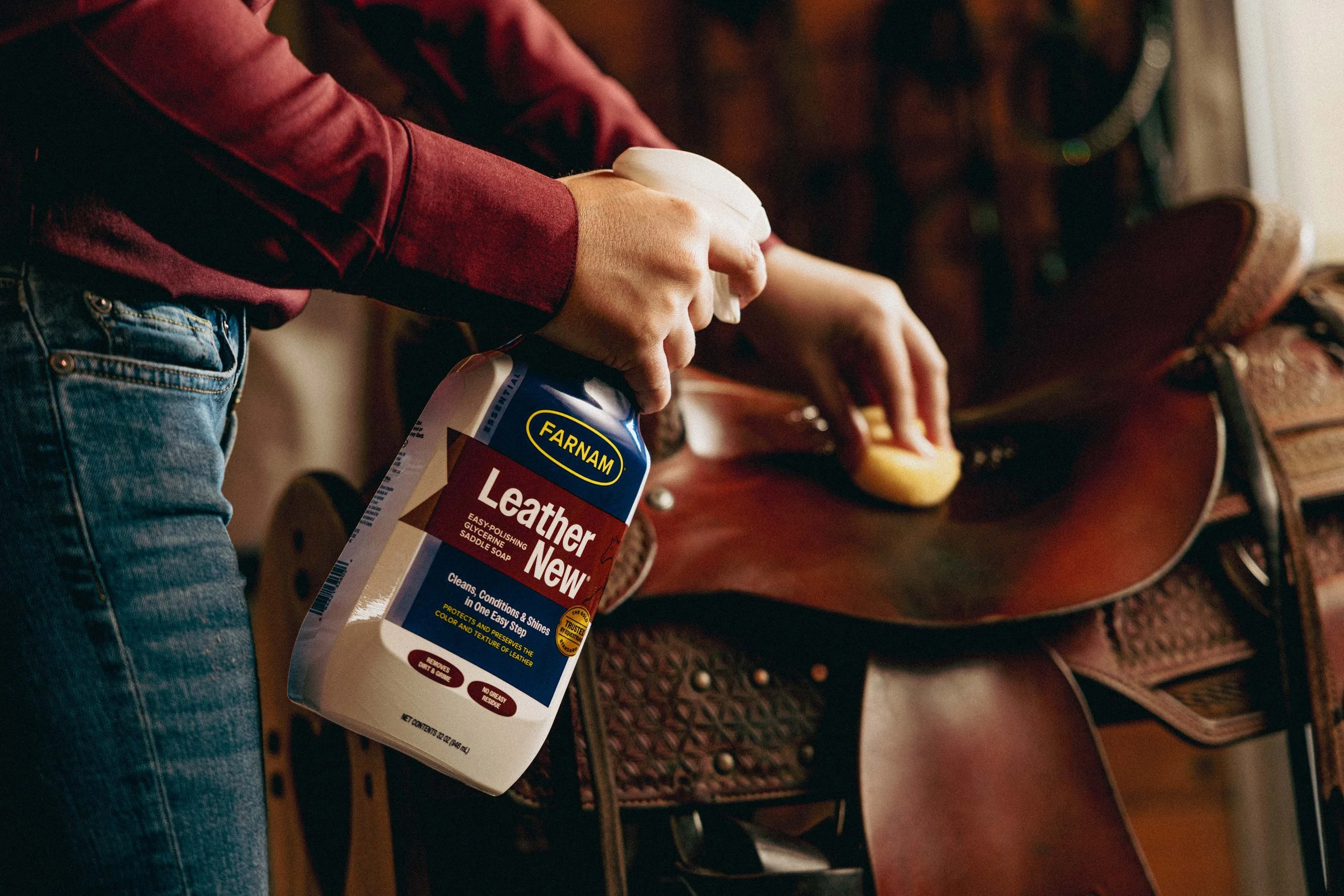 A person is cleaning a leather saddle with a spray bottle of leather cleaner and a cloth, while another person is wiping the saddle with a sponge.