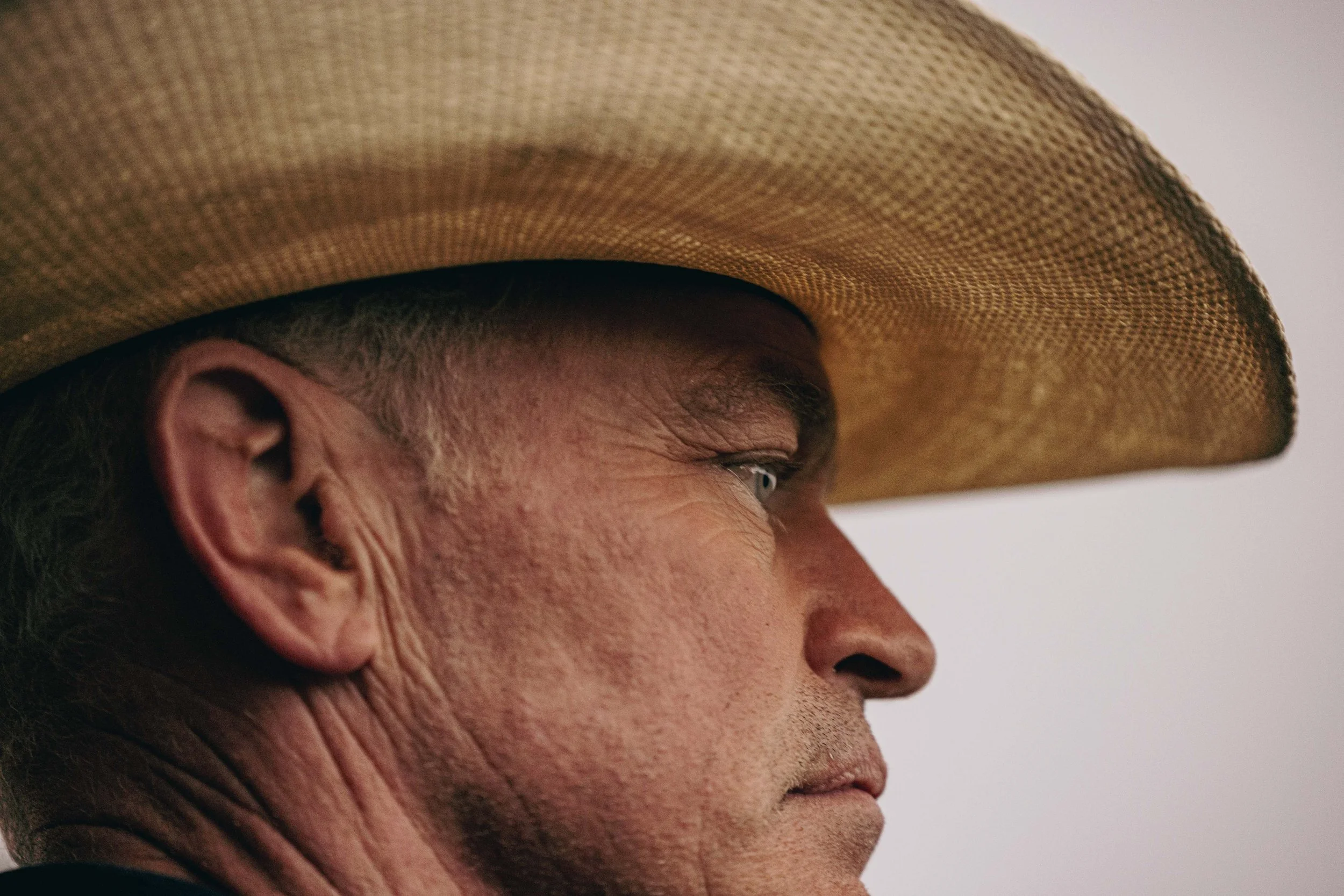 Close-up profile of a man wearing a large straw hat, showing his ear, eye, and part of his face with a neutral expression.
