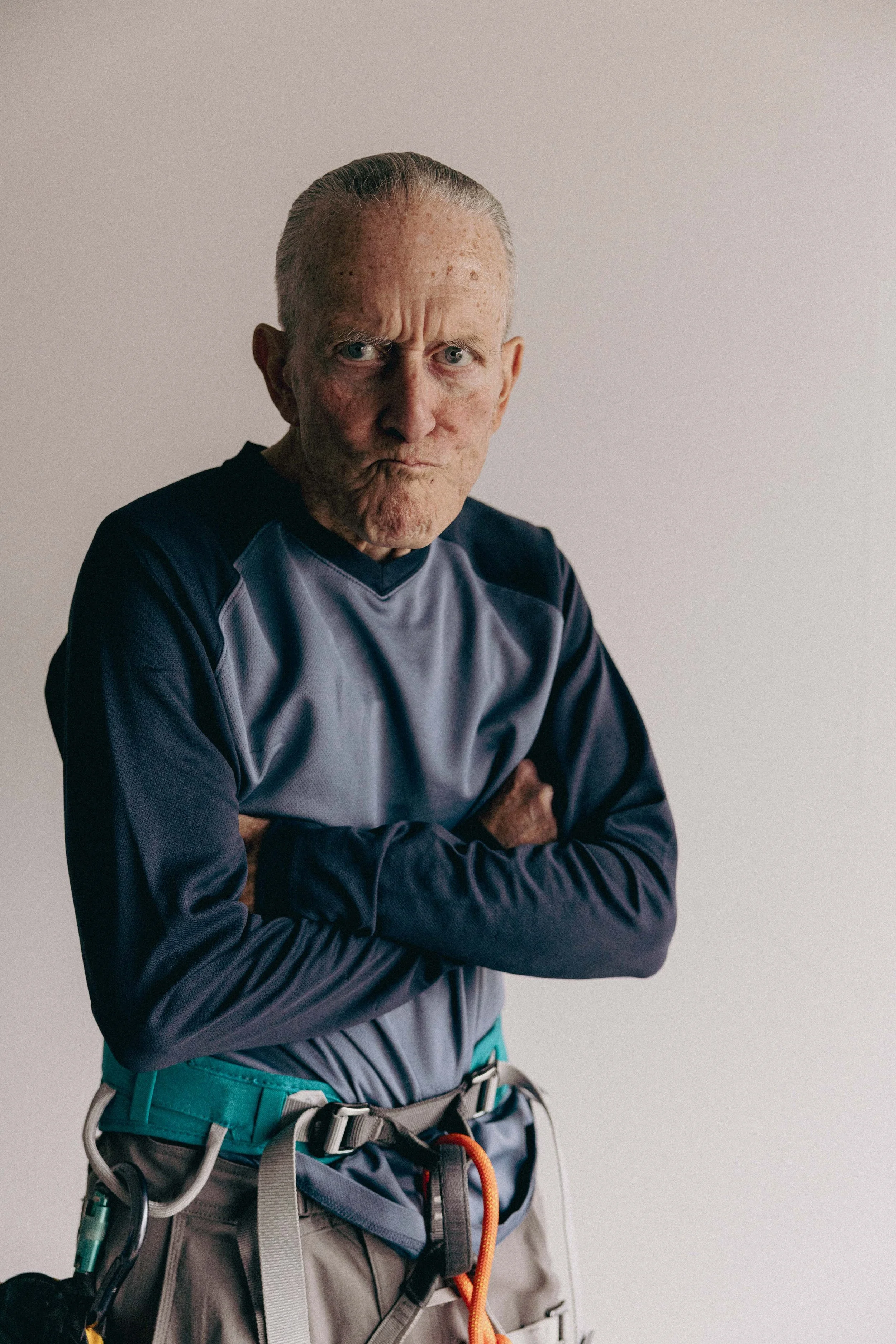 An elderly man with arms crossed, wearing a blue and gray shirt and climbing gear, making a stern face against a plain light background.