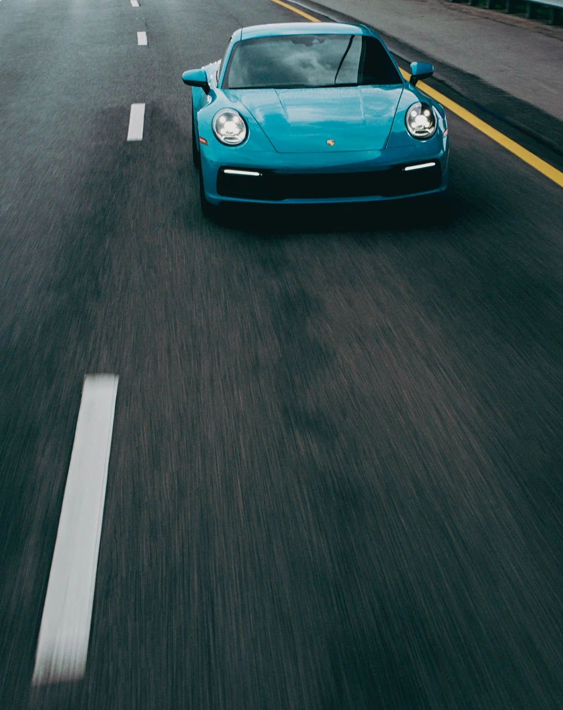 Blue sports car driving on a highway with lane markings and a yellow side barrier.