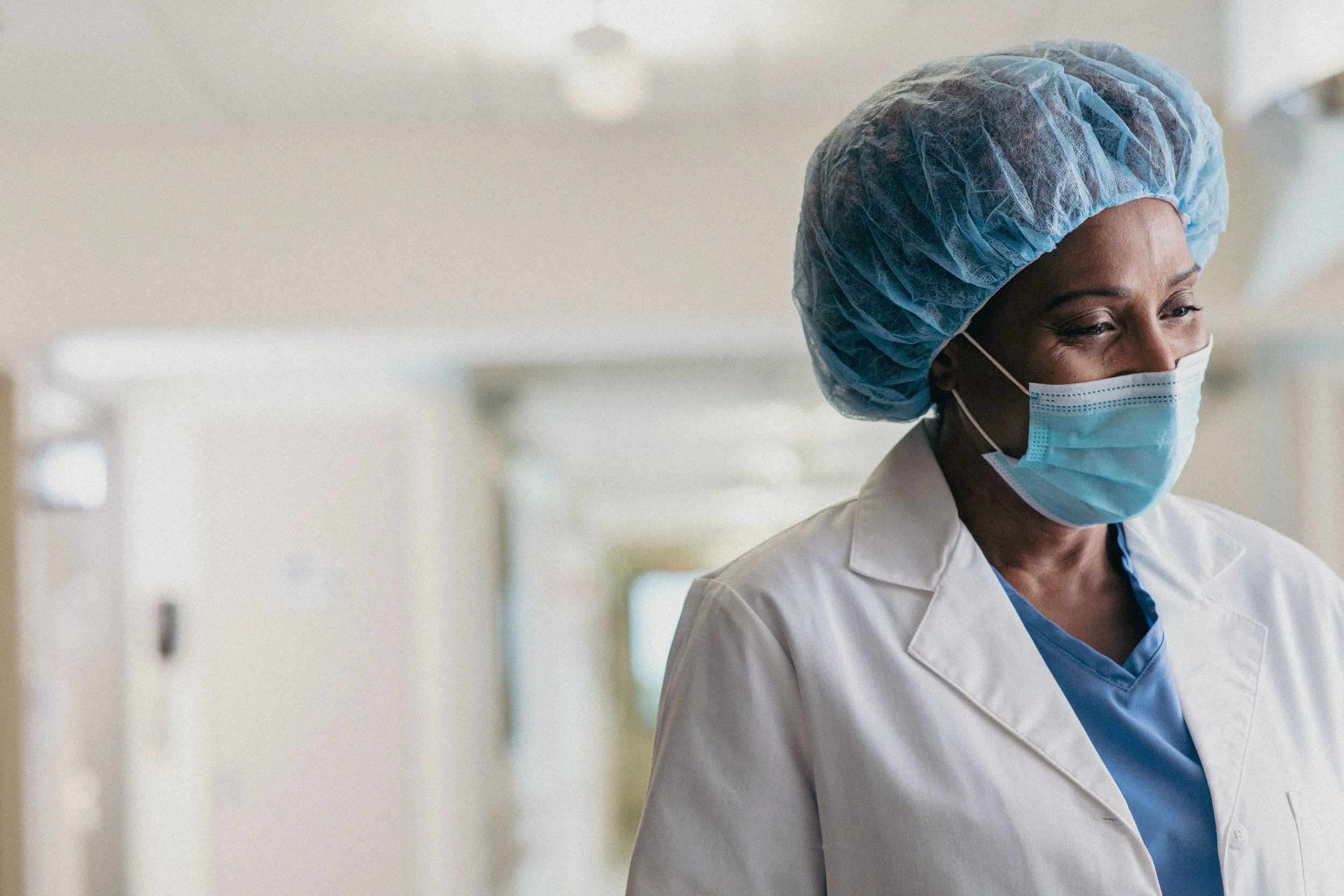 A healthcare worker wearing a blue surgical cap, a face mask, and a white lab coat in a medical facility.