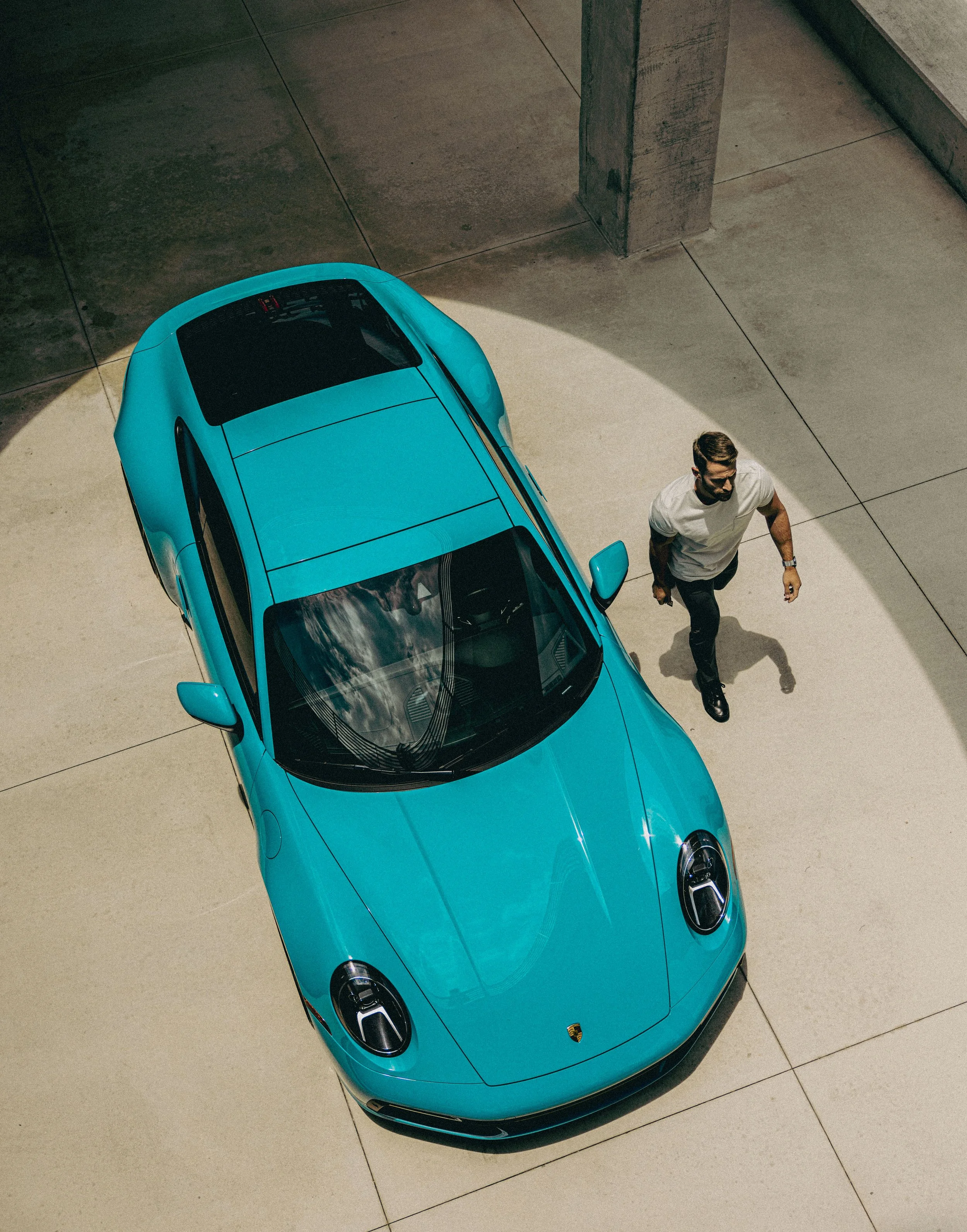 A top-down view of a vibrant blue sports car parked on a concrete surface near a building corner, with a man walking beside it.