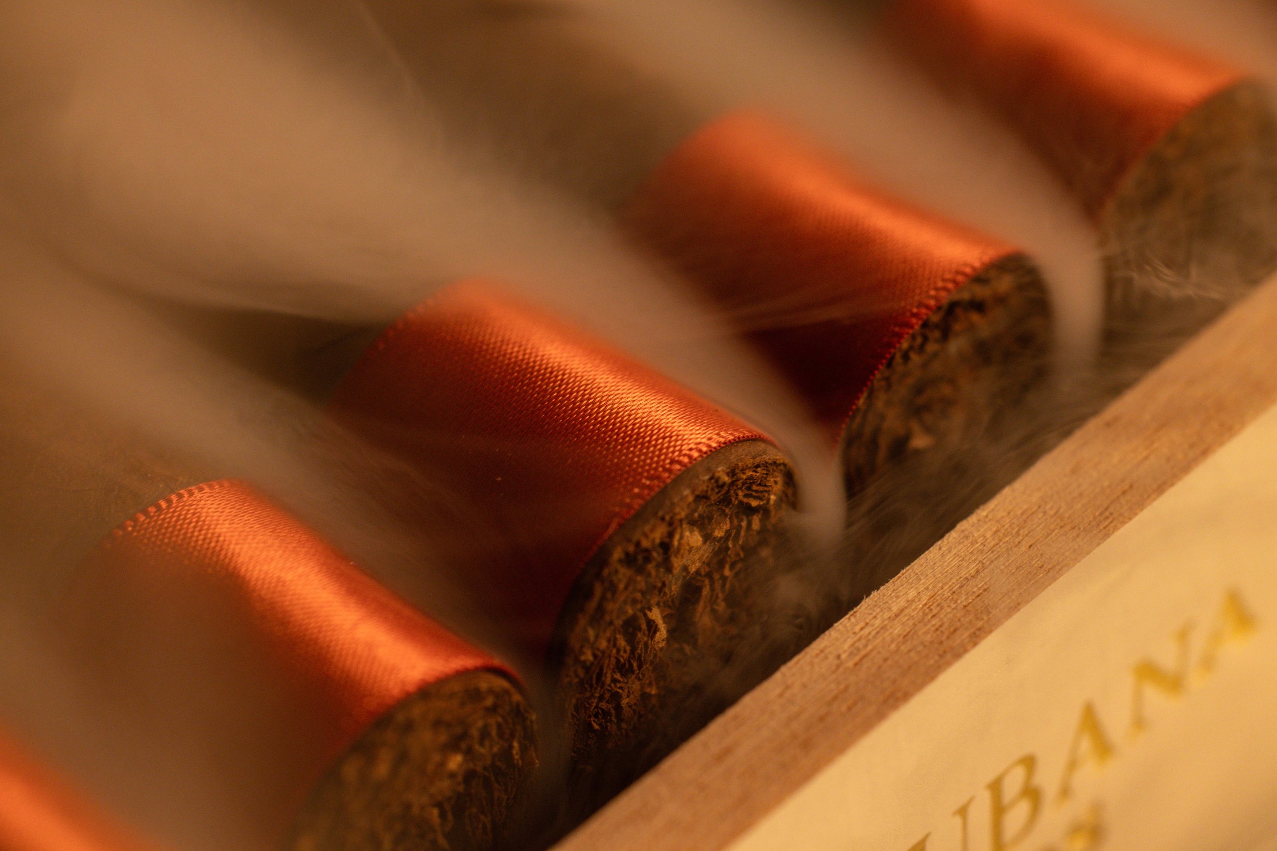 Close-up of a cigar box with several cigars inside, featuring orange-brown bands and smoke rising from them.