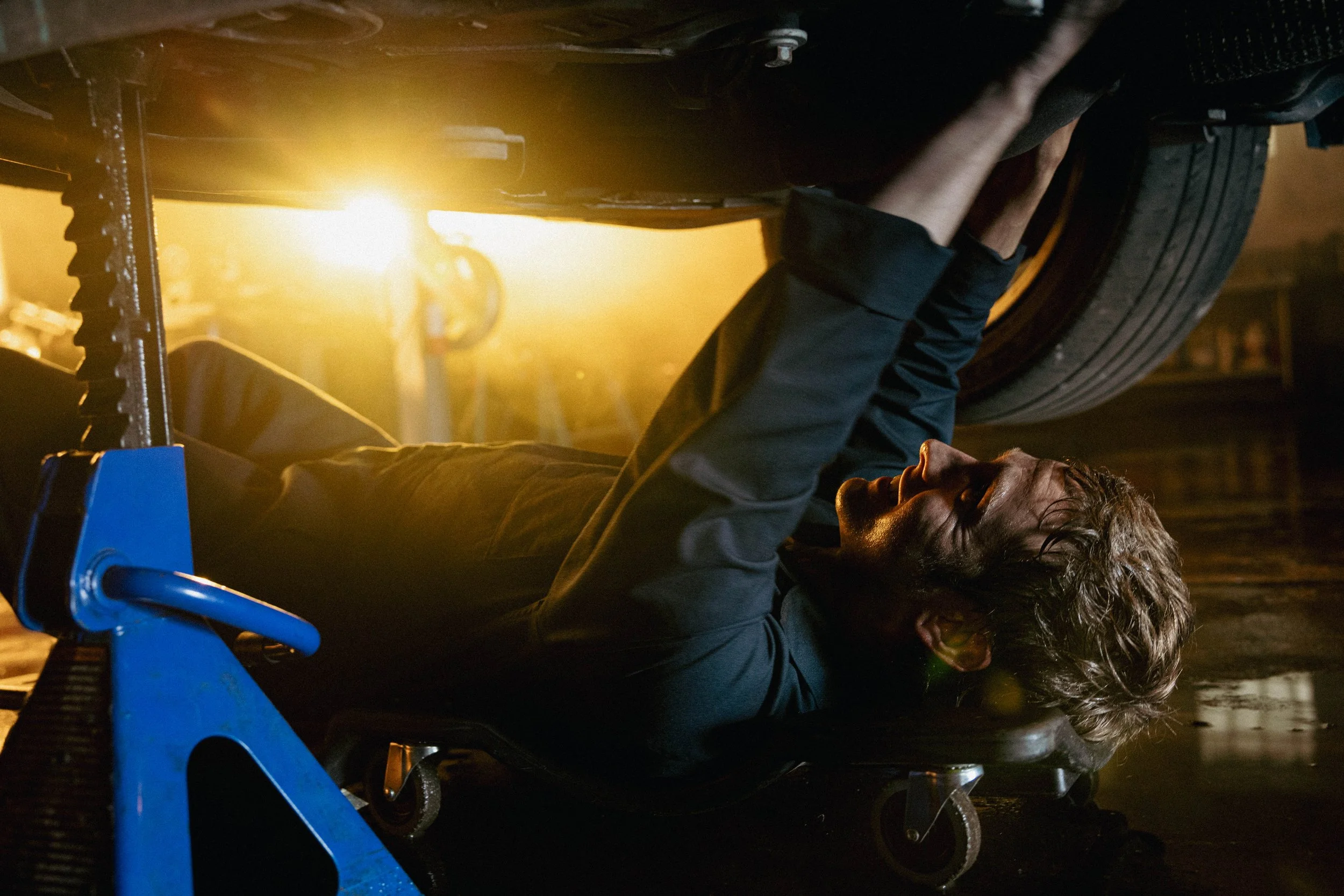 Man lying on a mechanic's creeper under a car, working on the underside of the vehicle in a garage with yellow light.