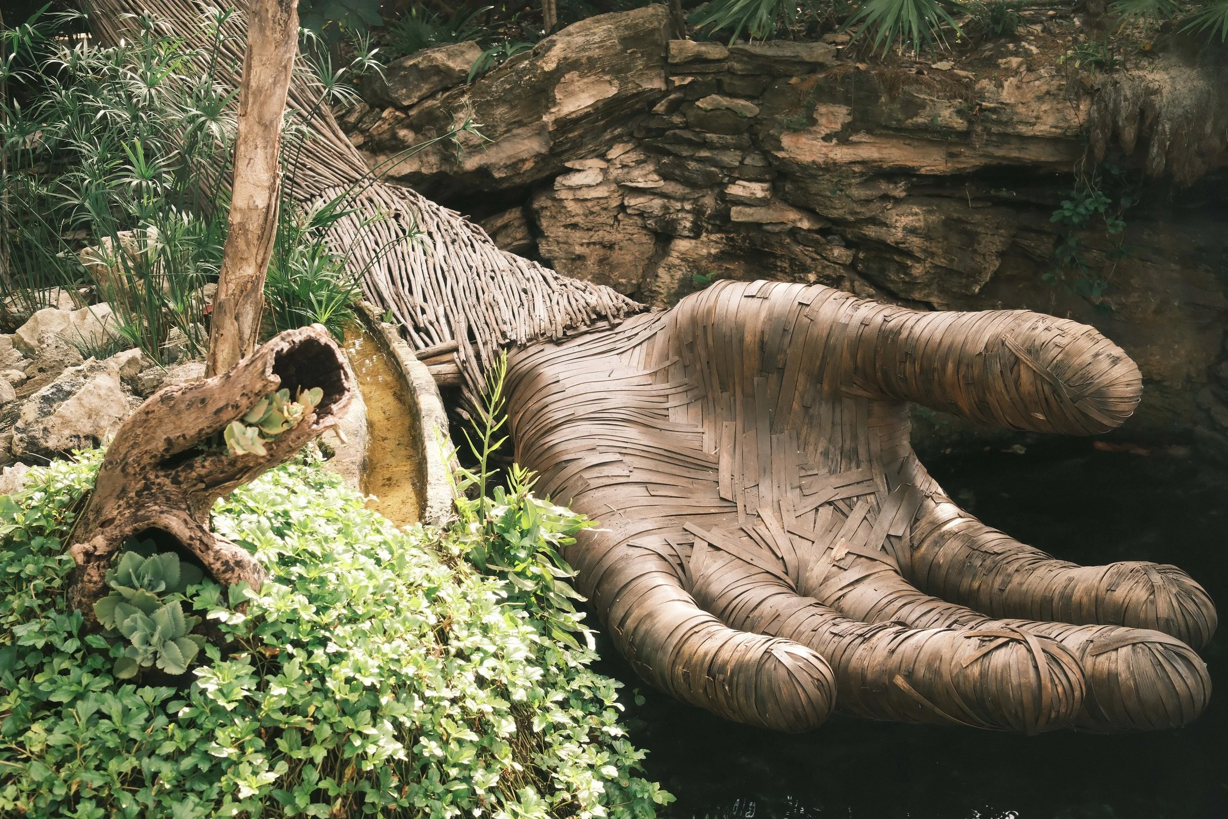 An artistic sculpture of a hand made from woven bamboo, situated among lush green plants and rocks, with a rocky background.
