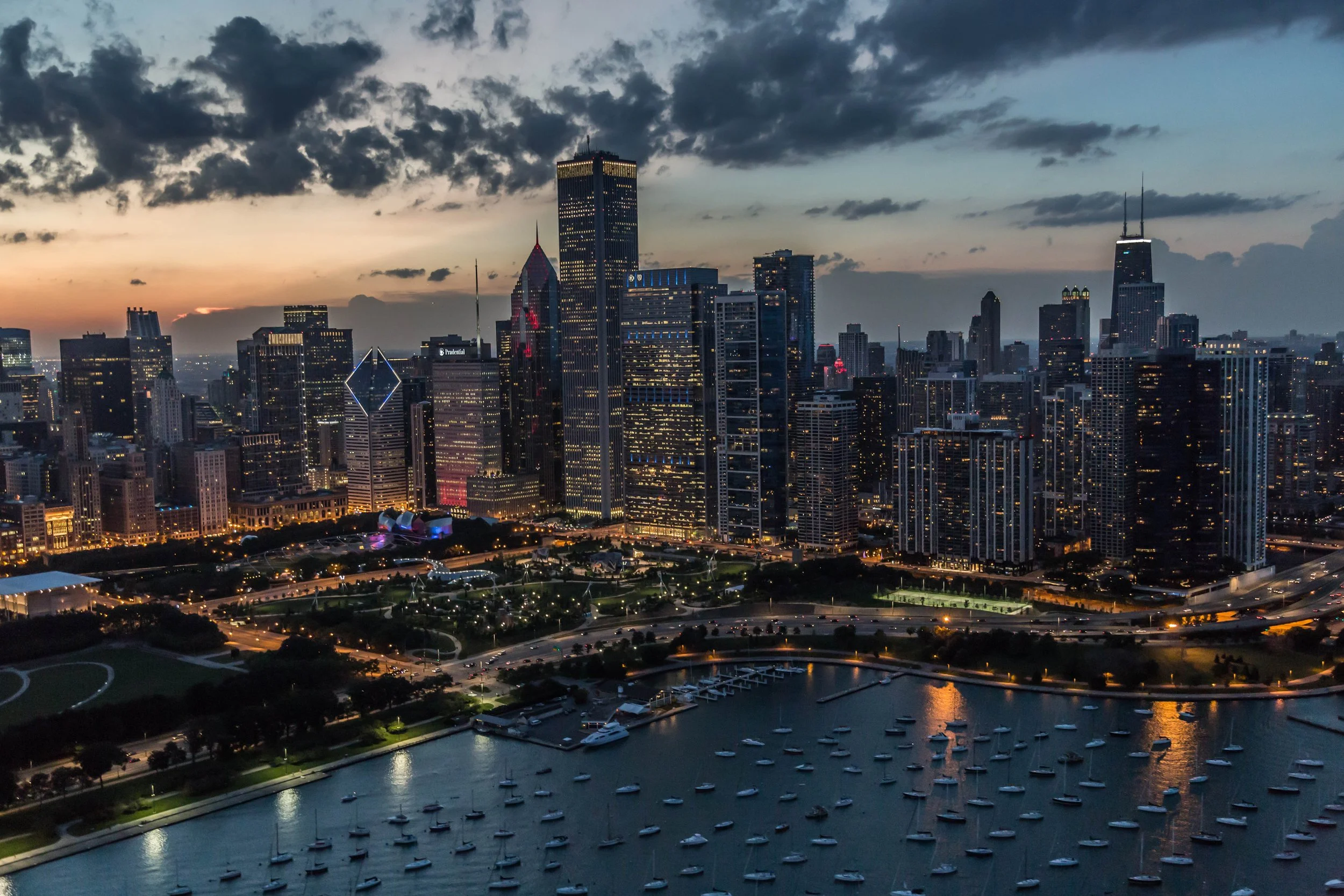 Aerial view of downtown Chicago at sunset with illuminated skyscrapers, a park, and boats in a harbor.