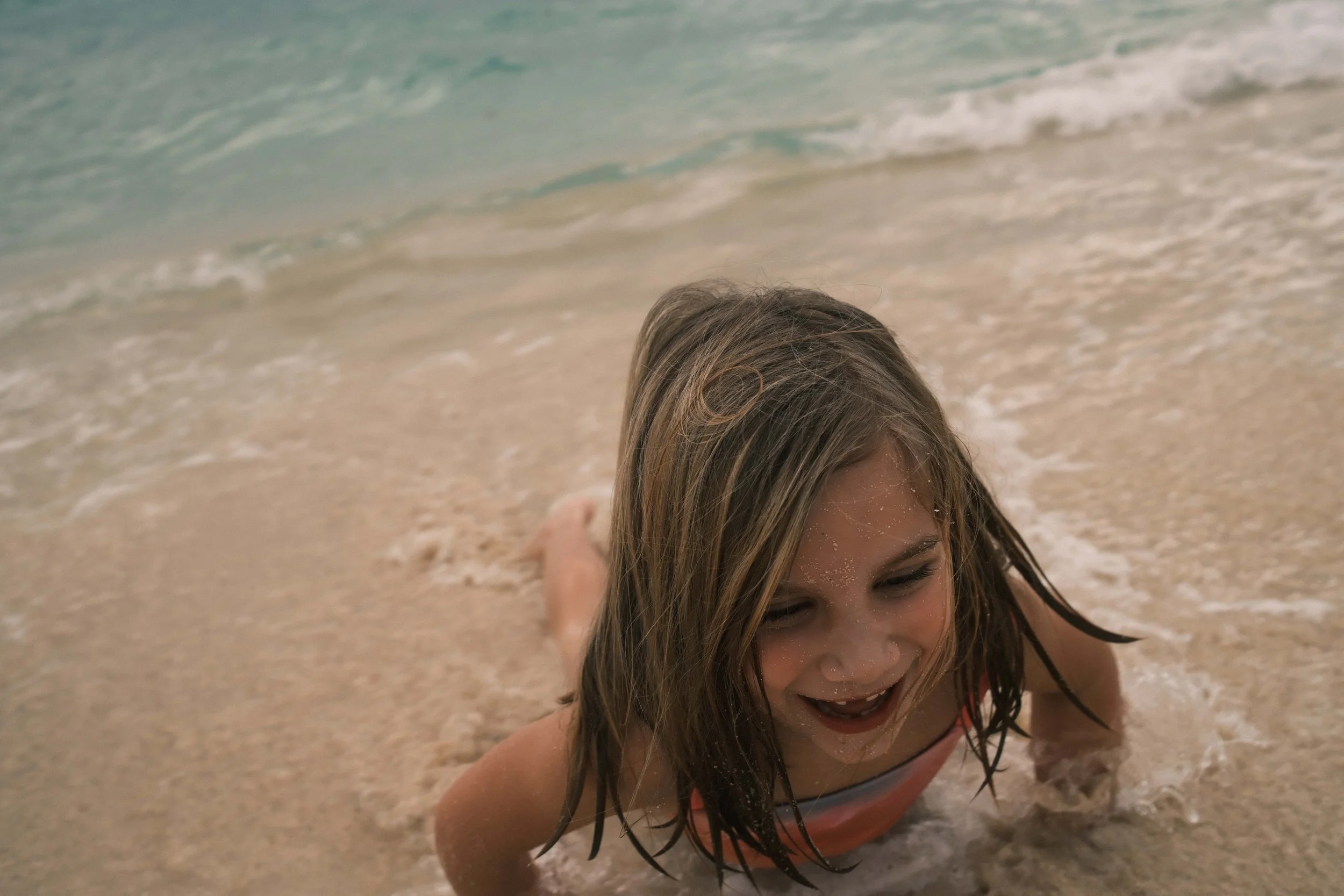 Young girl with wet hair smiling as she crawls on the sandy beach with ocean waves in the background.