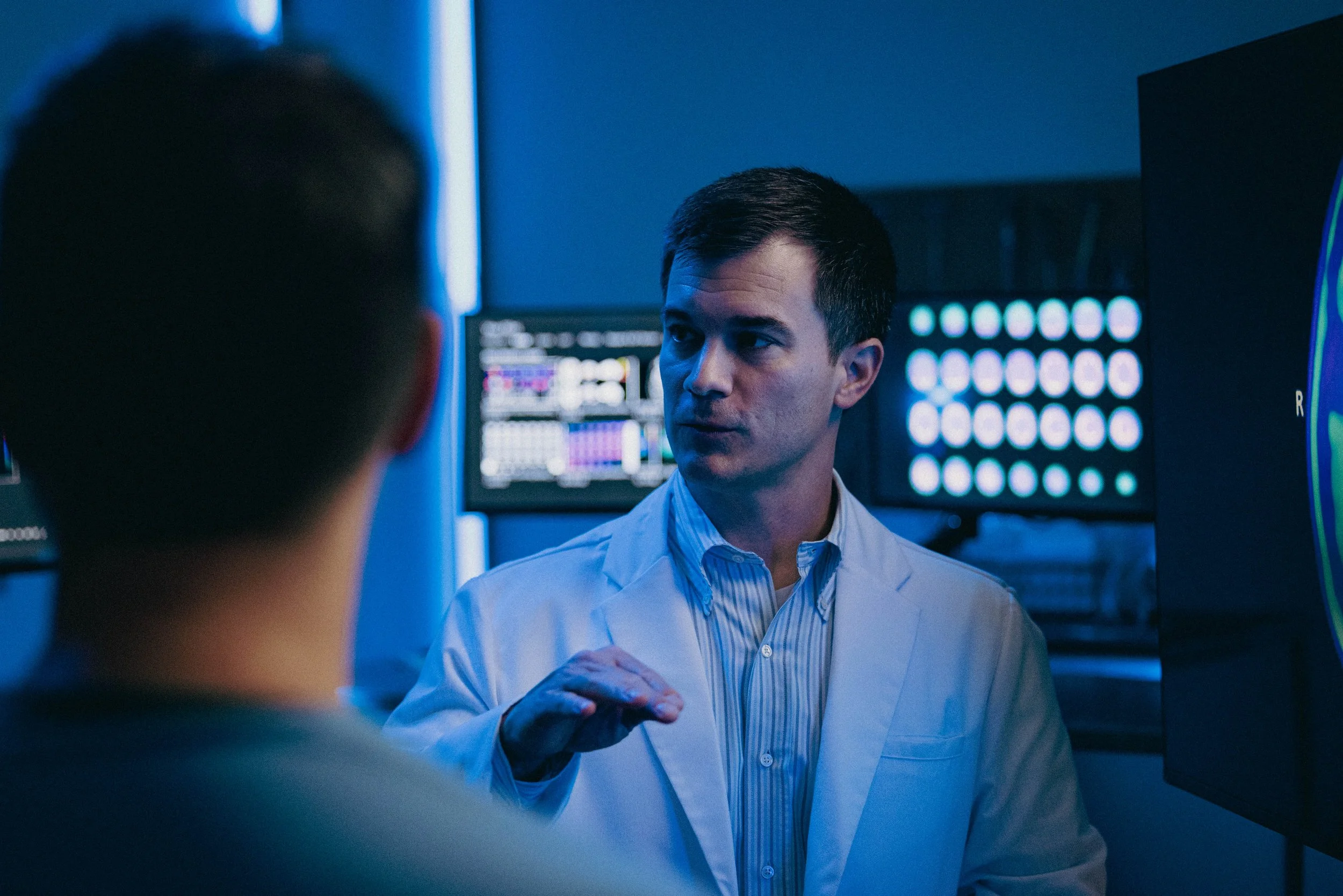 A male doctor in a white coat talking to a patient in a medical facility with monitors in the background.