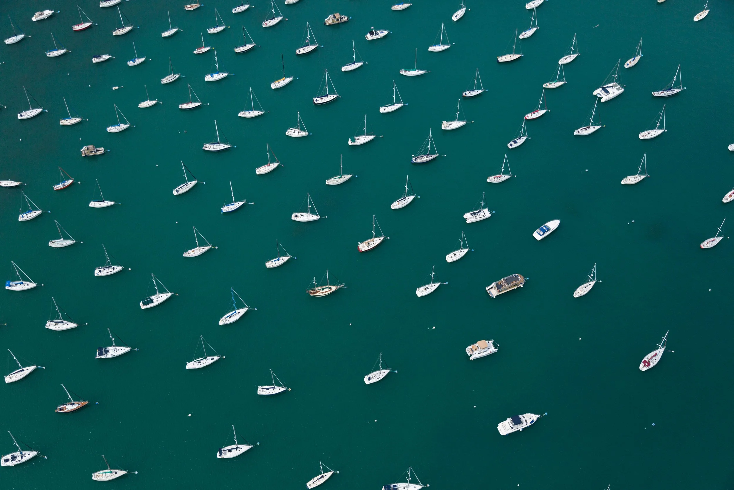 An aerial view of numerous sailboats and a couple of motorboats anchored in a marina or harbor with teal-green water.
