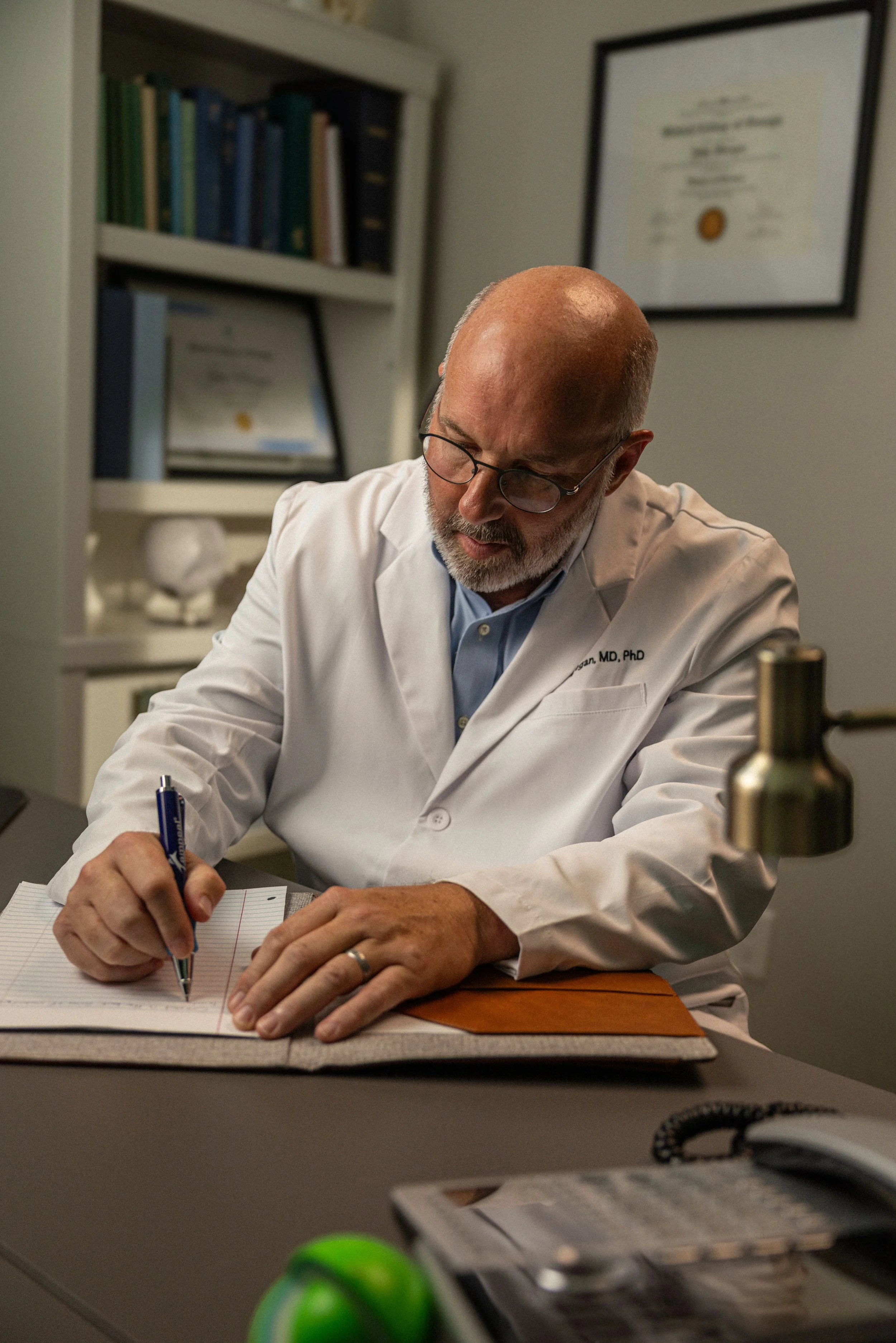 A male doctor with glasses and a beard, wearing a white lab coat, is sitting at a desk and writing in a notebook.