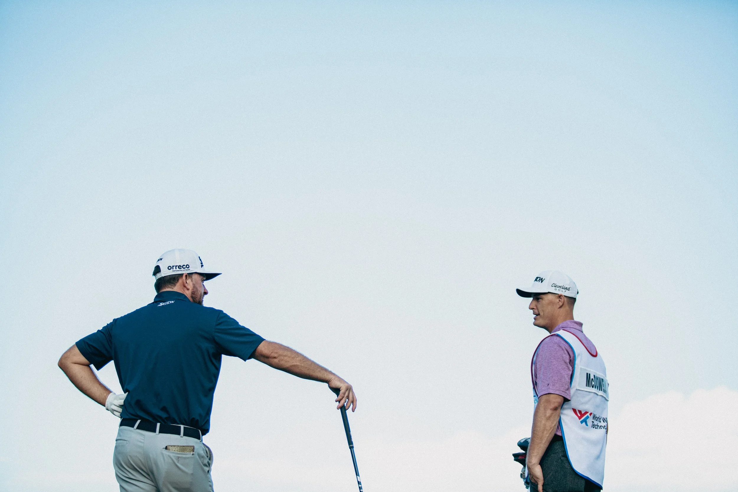 Two men on a golf course, one in a blue polo shirt and beige pants with a golf club, and the other in a cap and a golf vest, engaging in conversation against a light blue sky.