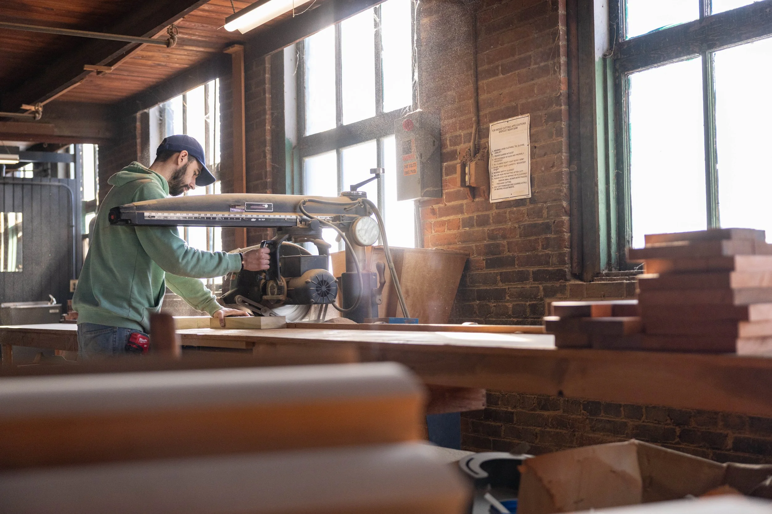 A man in a green hoodie and cap using a circular saw to cut wood in a woodworking shop with large windows and brick walls.