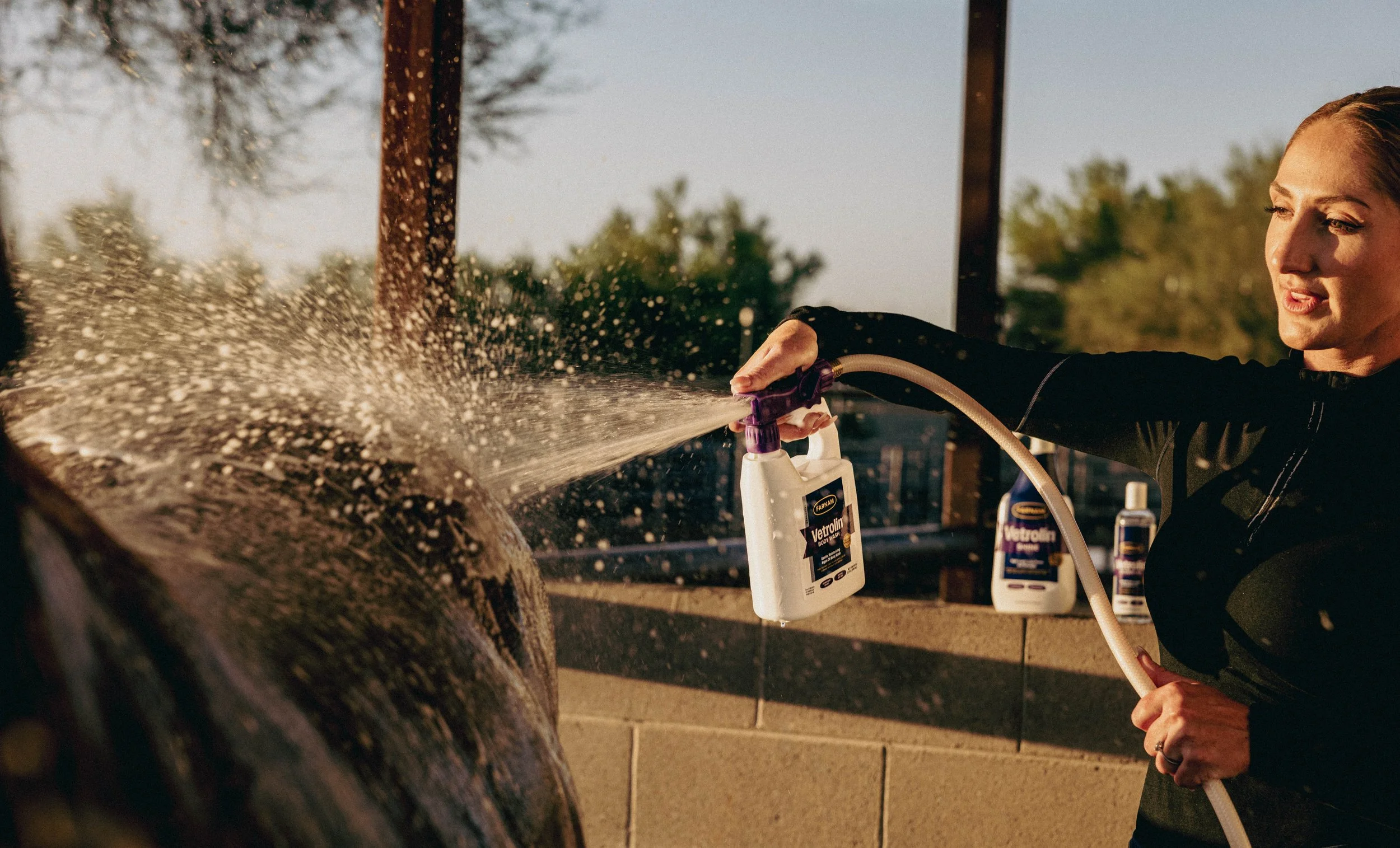 A woman spray washing a large rock with a garden hose outside during daytime, with bottles of cleaning product in the background.