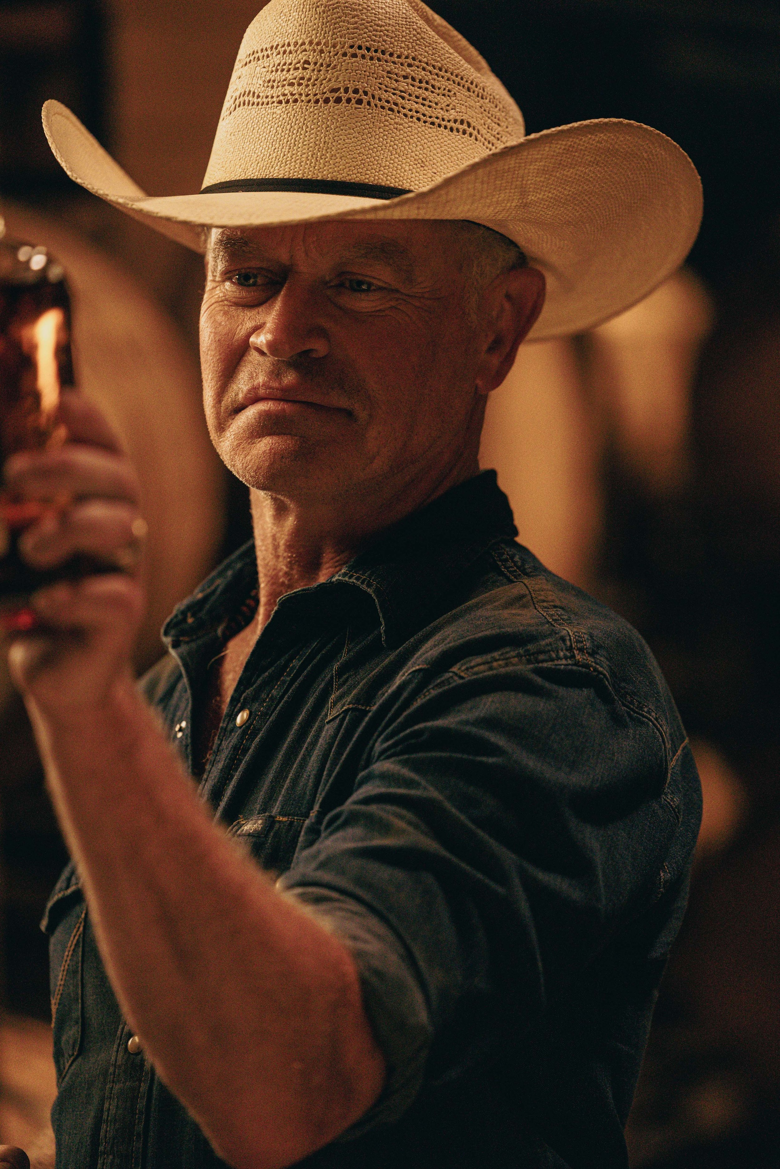 A man wearing a white cowboy hat and a dark denim shirt, holding a drink, in a dimly lit setting.