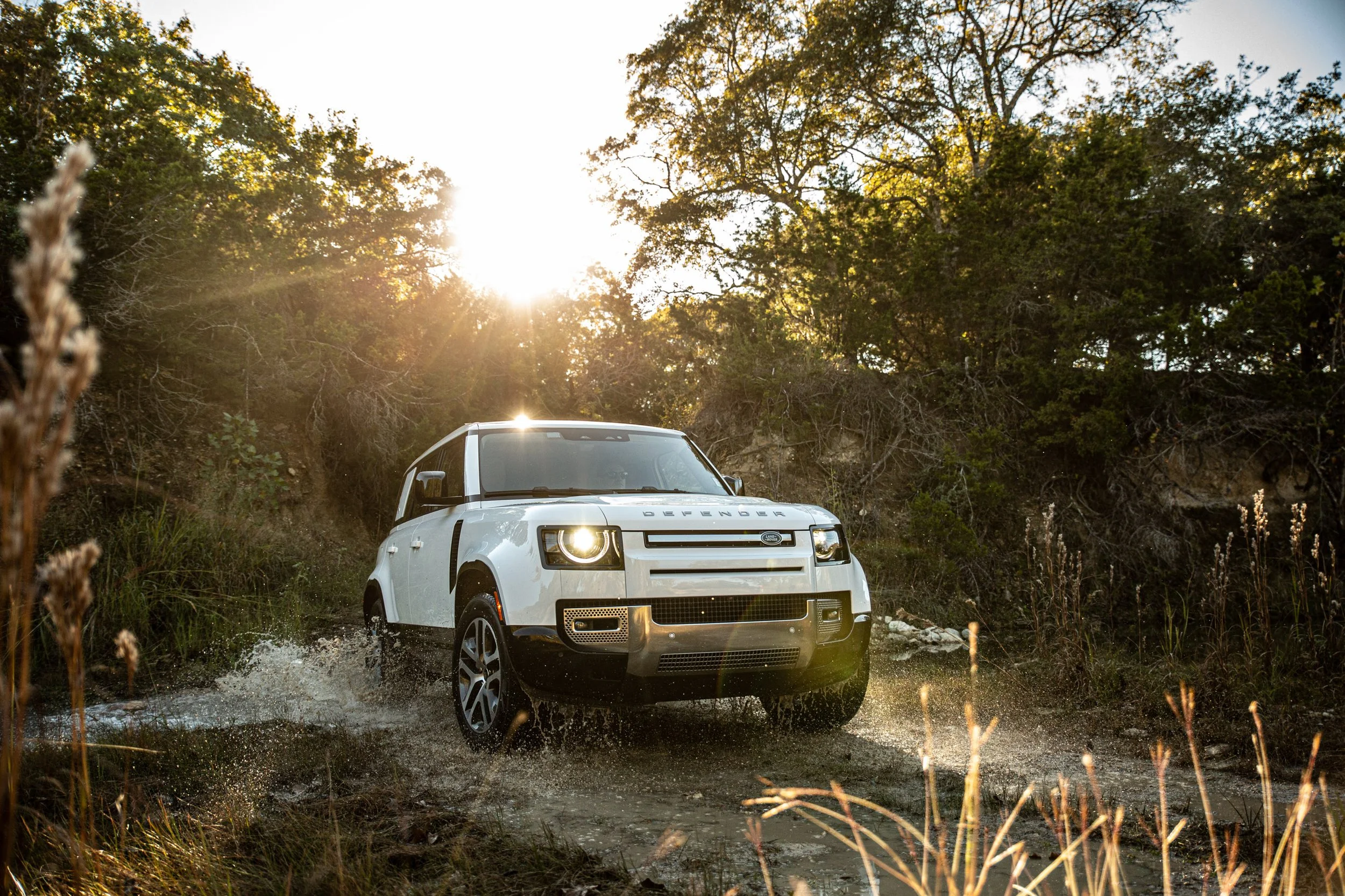 A white Land Rover Defender driving through a muddy trail in a forested area with sunlight shining through trees in the background.