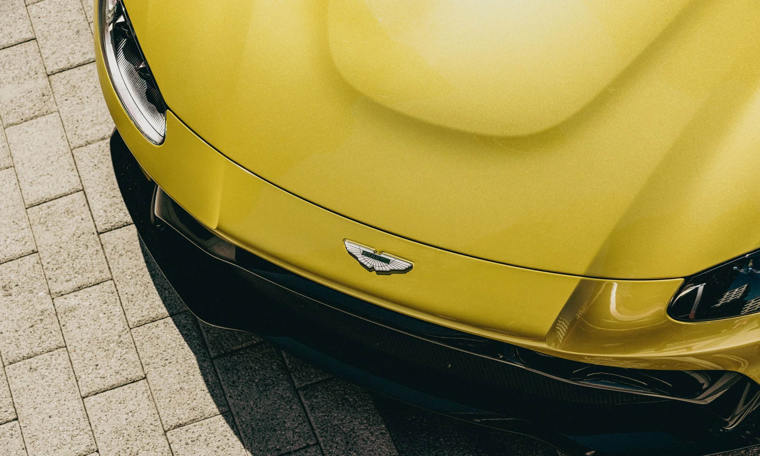 Close-up of a yellow luxury sports car with an Aston Martin logo, parked on a paved surface.