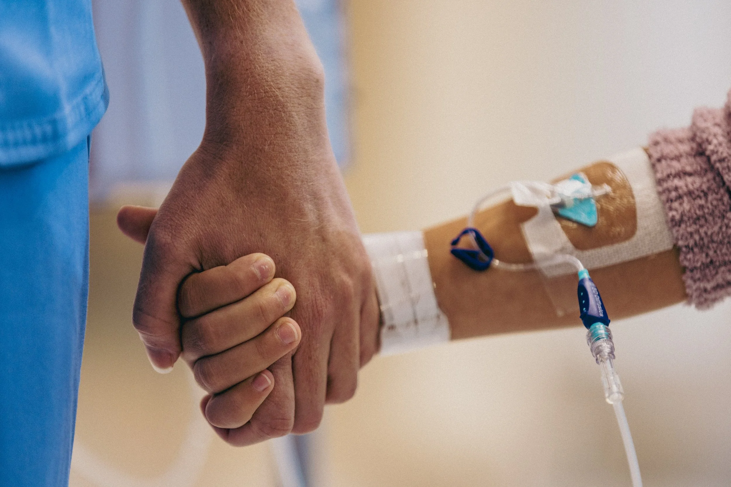 Close-up of two hands holding, one with a hospital IV catheter inserted and taped to the arm.