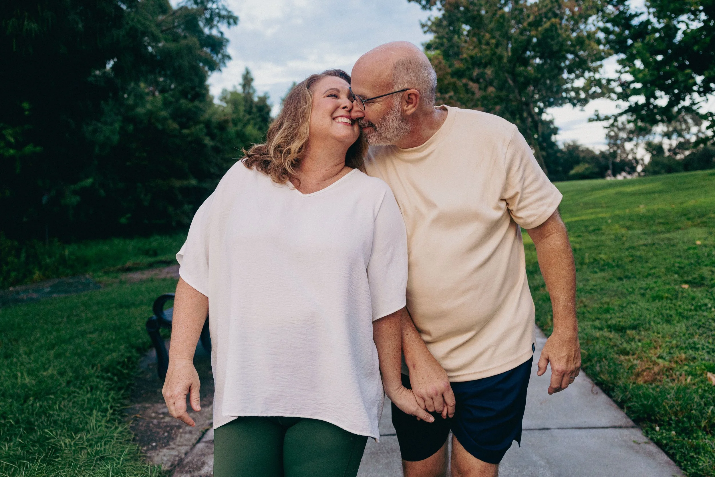 An elderly couple walking outdoors on a park pathway, smiling and holding hands, with trees and grass in the background.