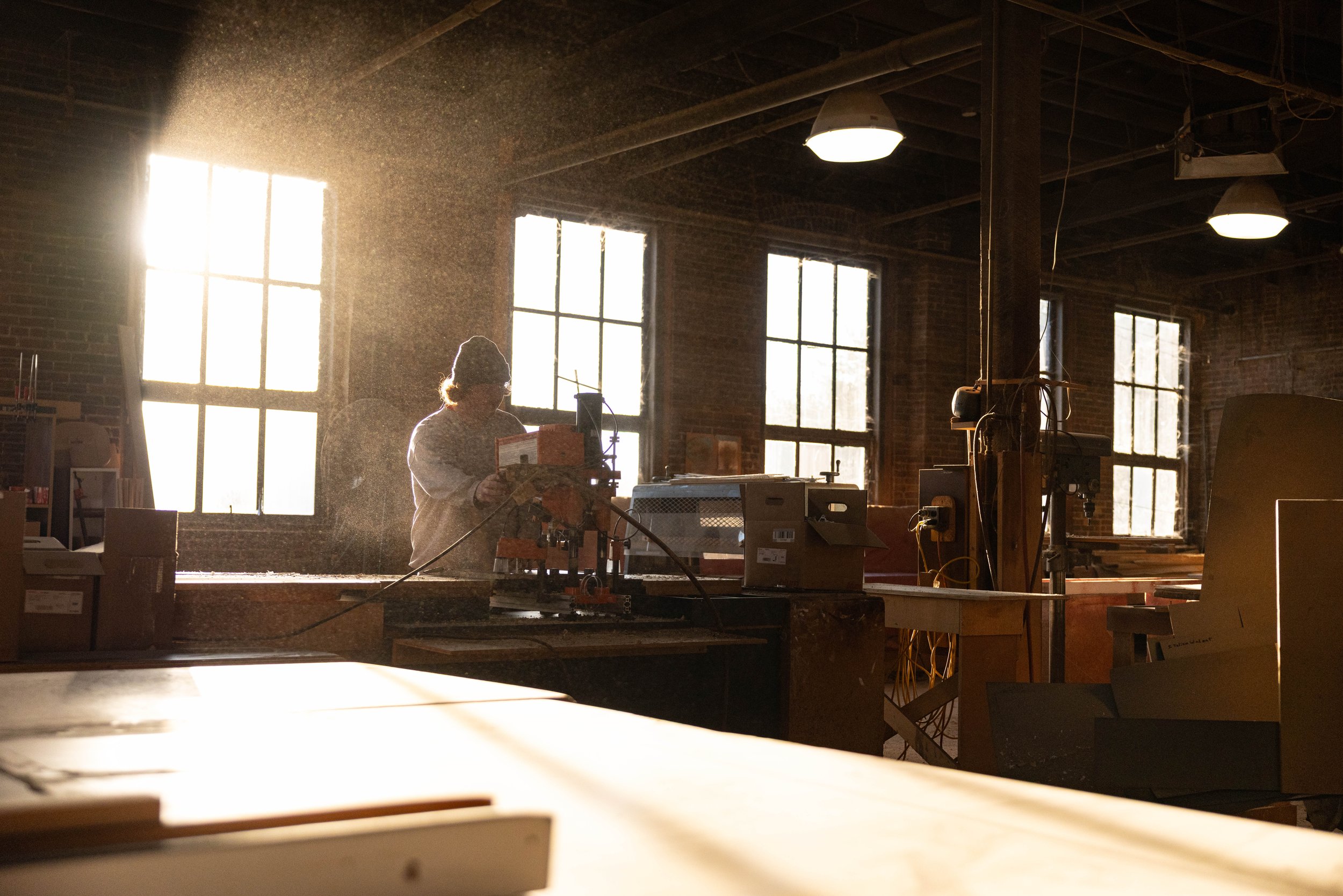 Person working in a woodworking workshop with sunlight streaming through large windows.
